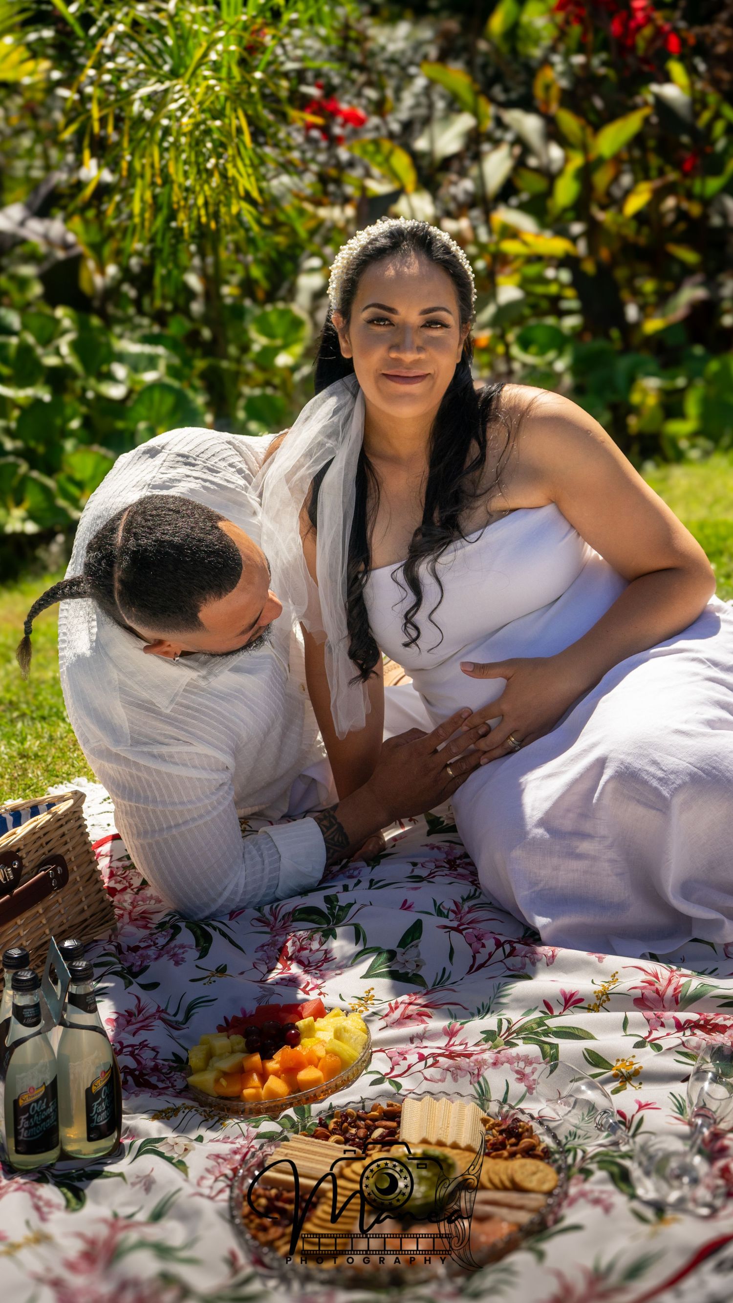 Pregnant woman in white dress with partner kissing belly during outdoor garden picnic.