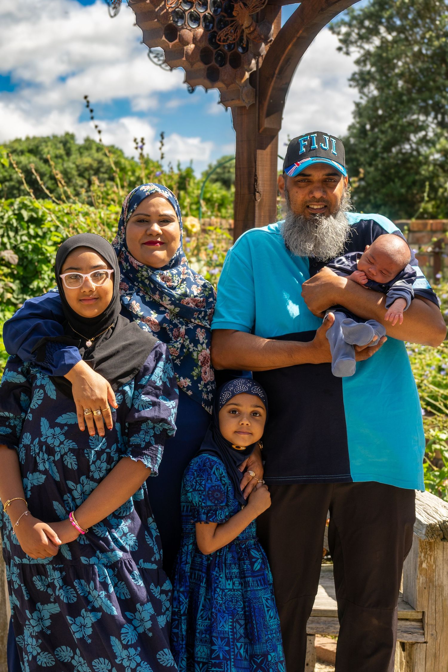 A Muslim family of five poses under a wooden arch in a sunny garden.