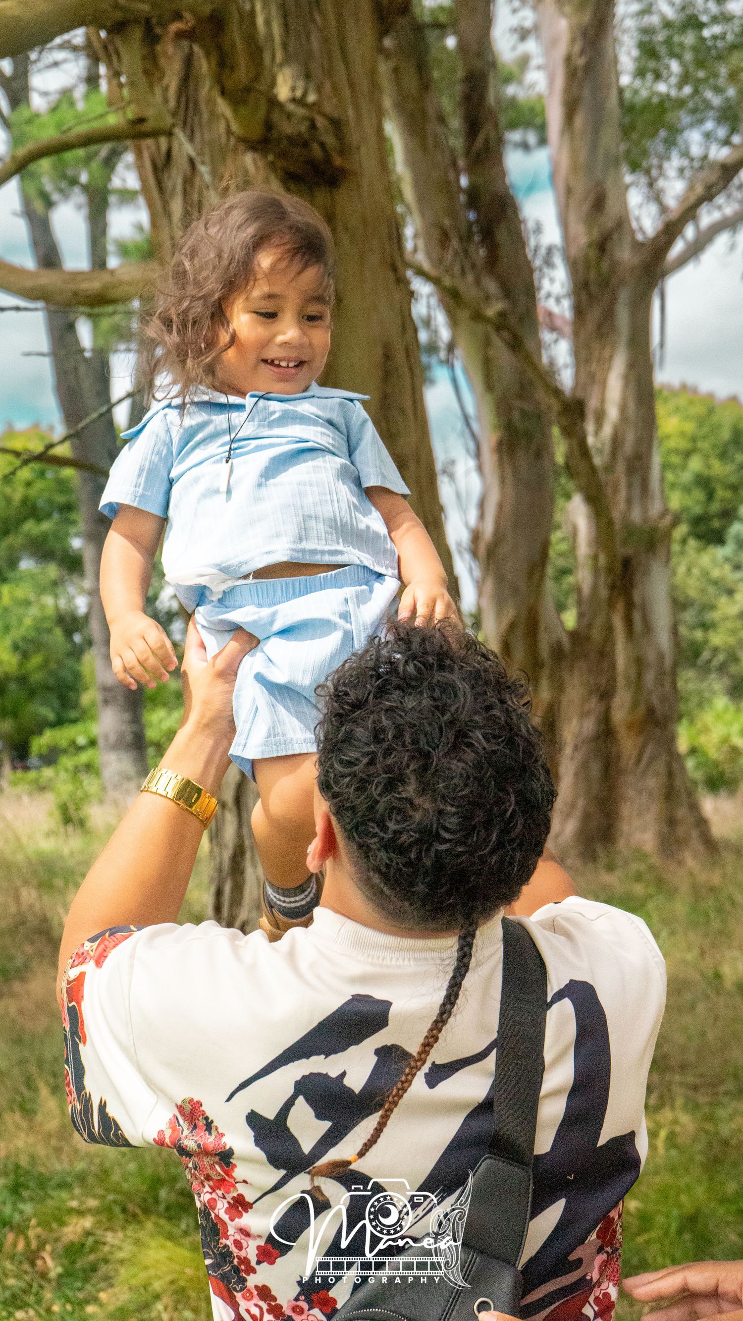 Father lifting happy toddler girl in blue outfit high into the air near large trees outdoors.