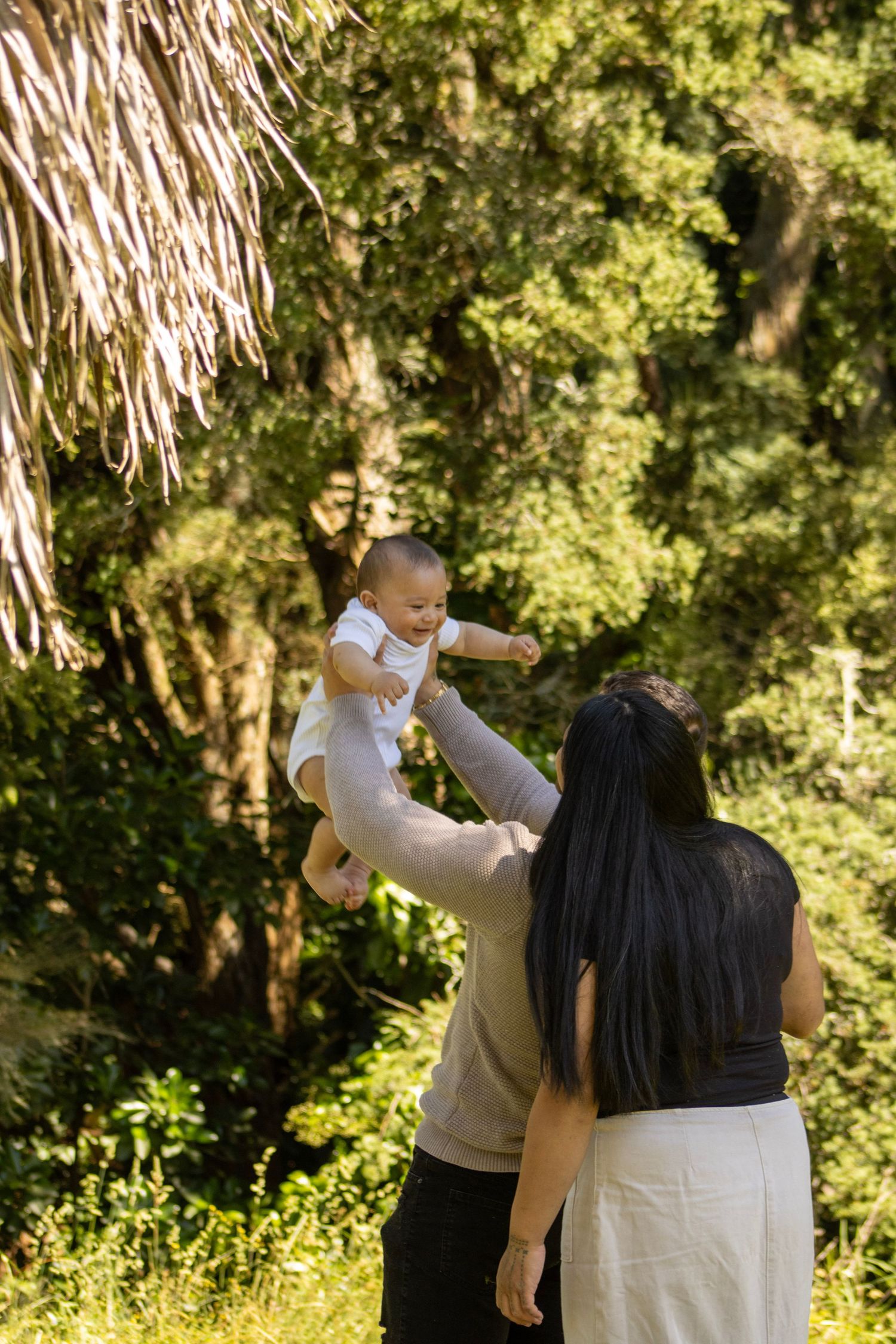 Parents joyfully lift their baby in the air outdoors surrounded by lush green trees.