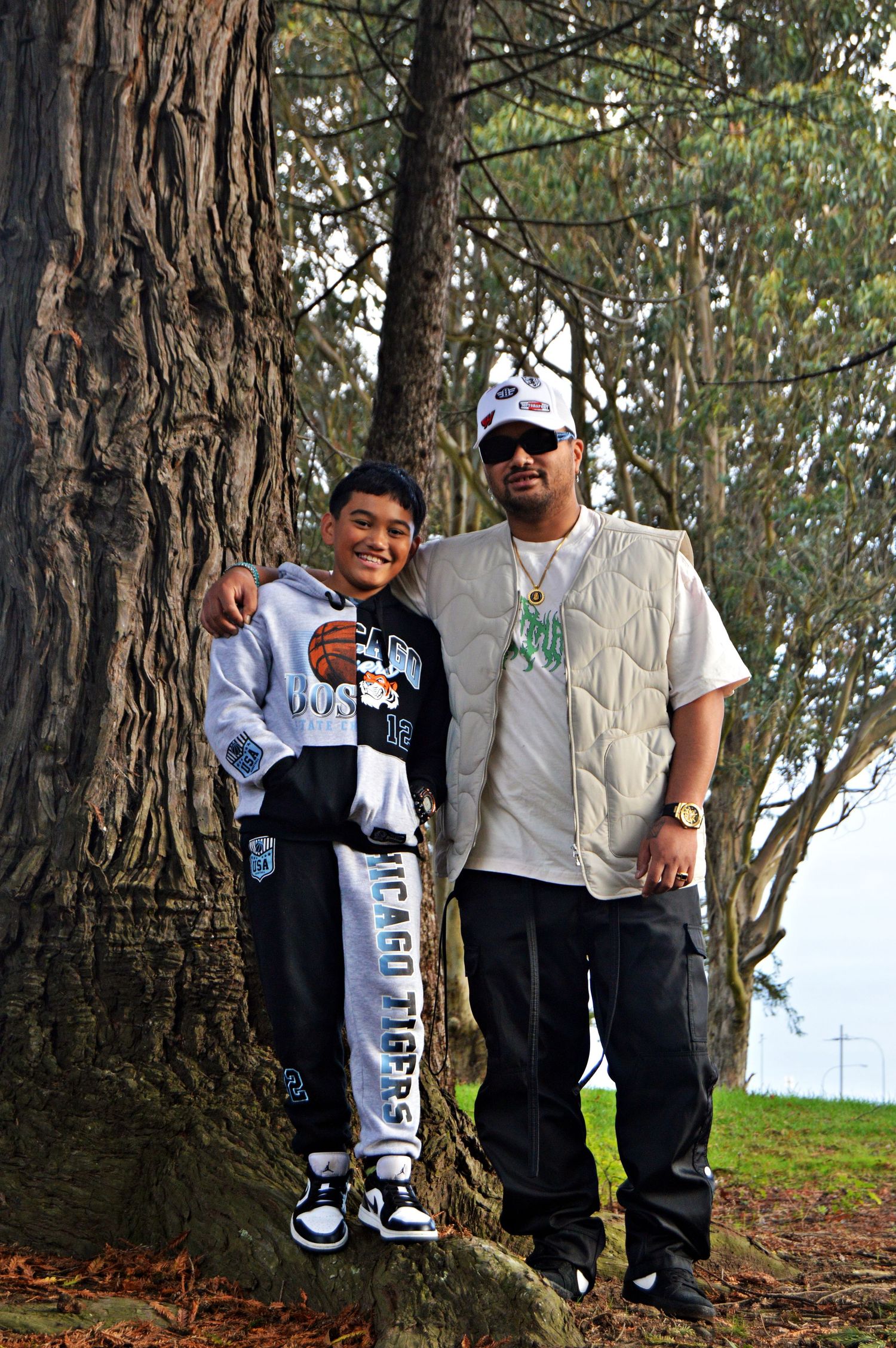 A father and son pose together against a large tree trunk in a park setting.