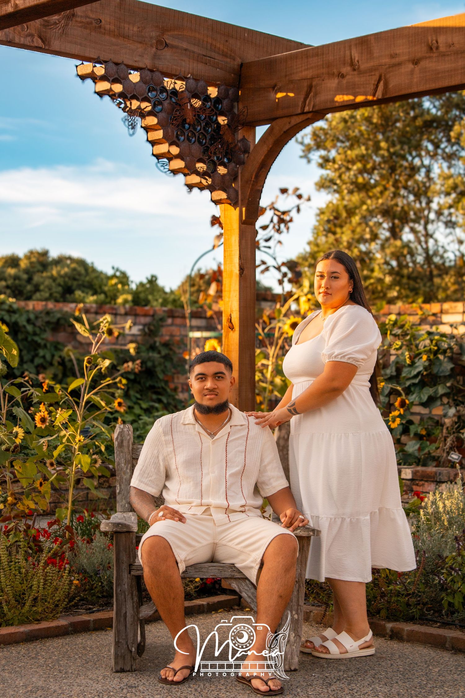 A couple in white outfits poses under a wooden pergola surrounded by sunflowers at golden hour.
