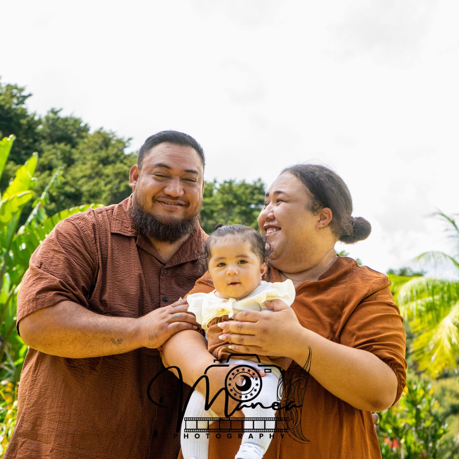 A smiling couple in brown outfits holds their baby outdoors with tropical foliage.