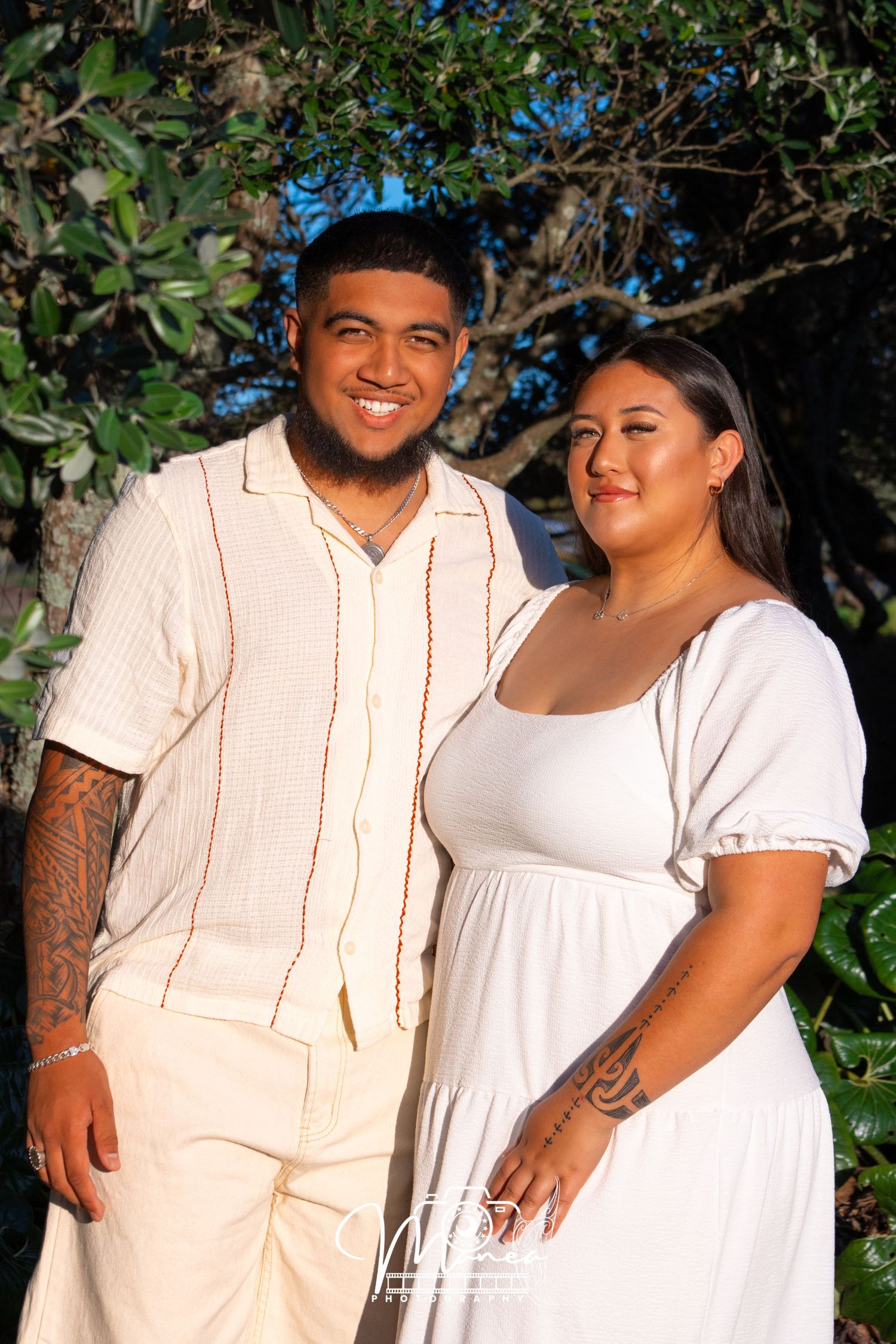 Smiling couple in coordinating cream and white outfits posing together outdoors under green trees.