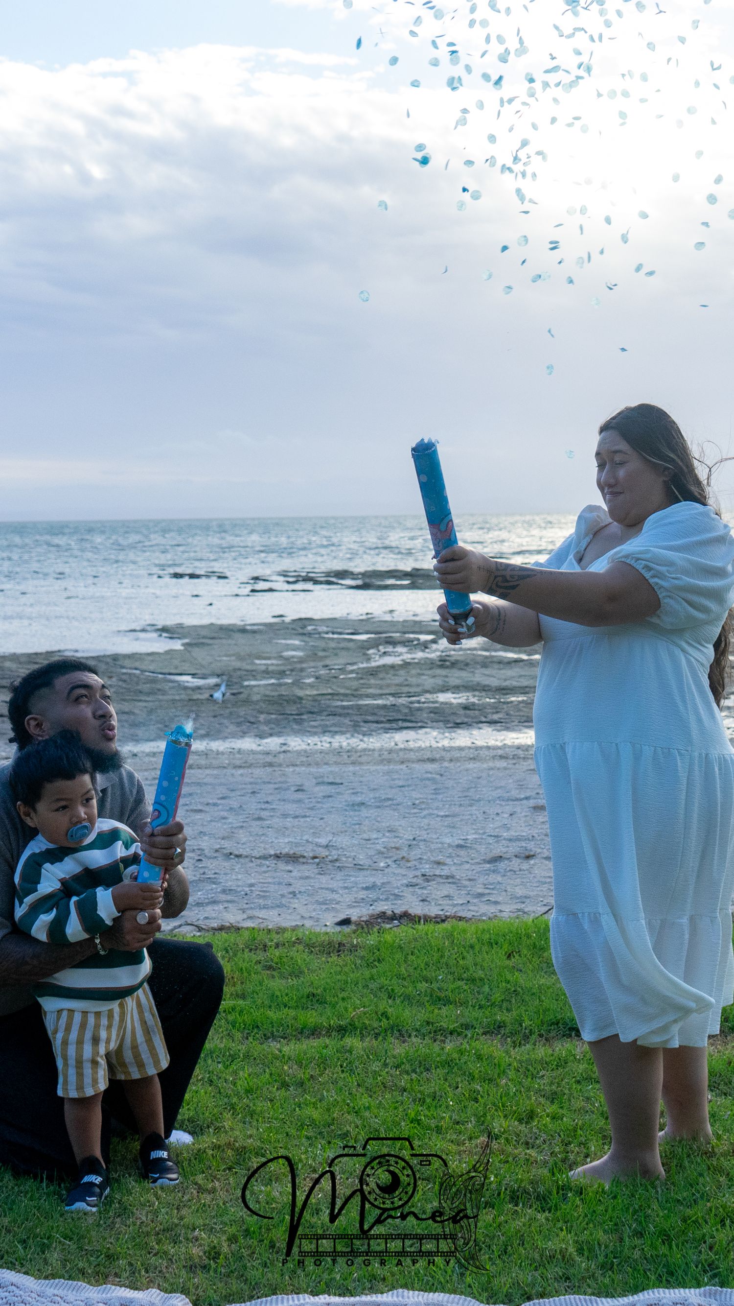 Family doing blue confetti gender reveal by the ocean on a cloudy day.