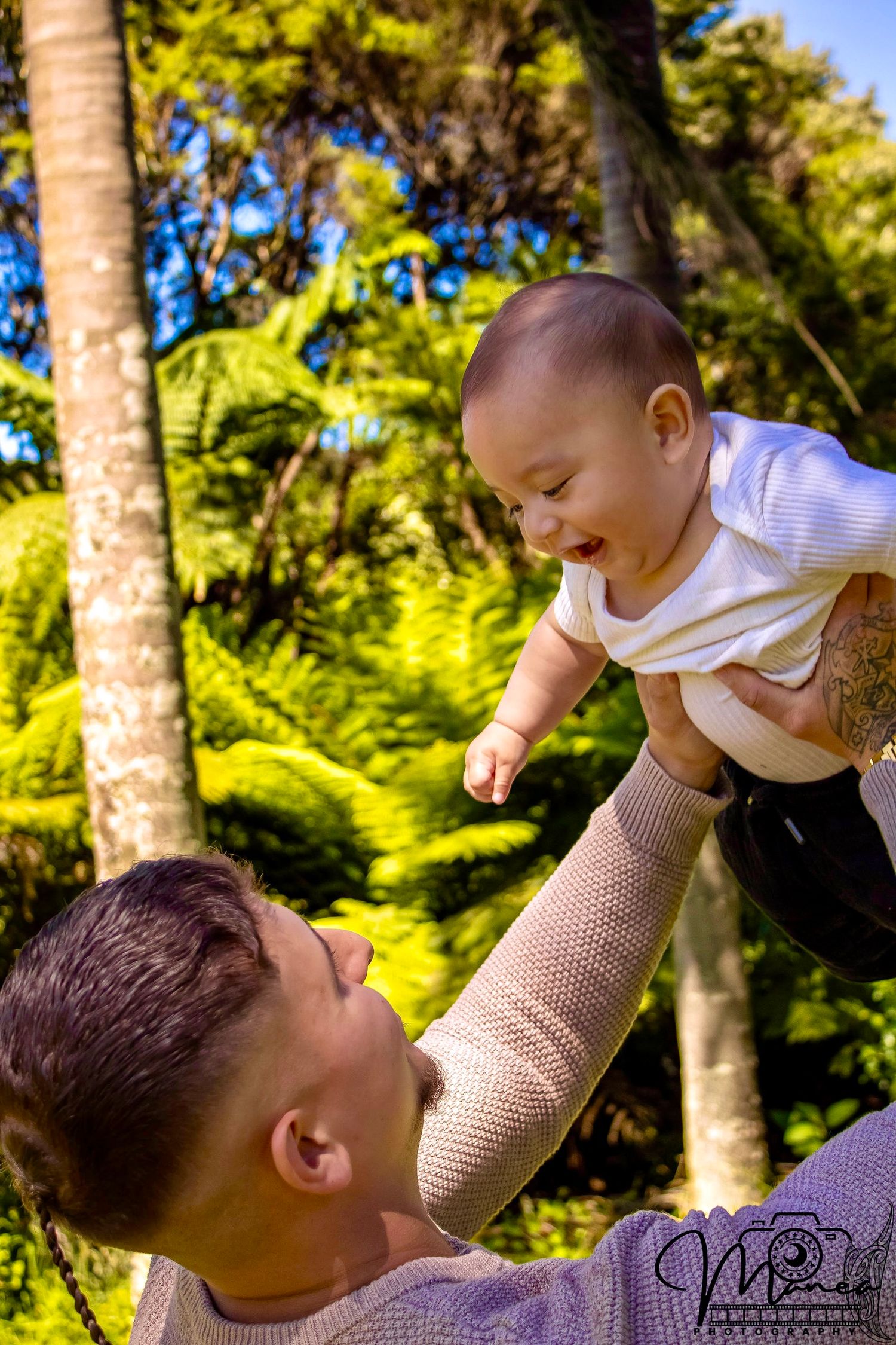 Joyful father lifting laughing baby high in the air against a lush tropical garden background.
