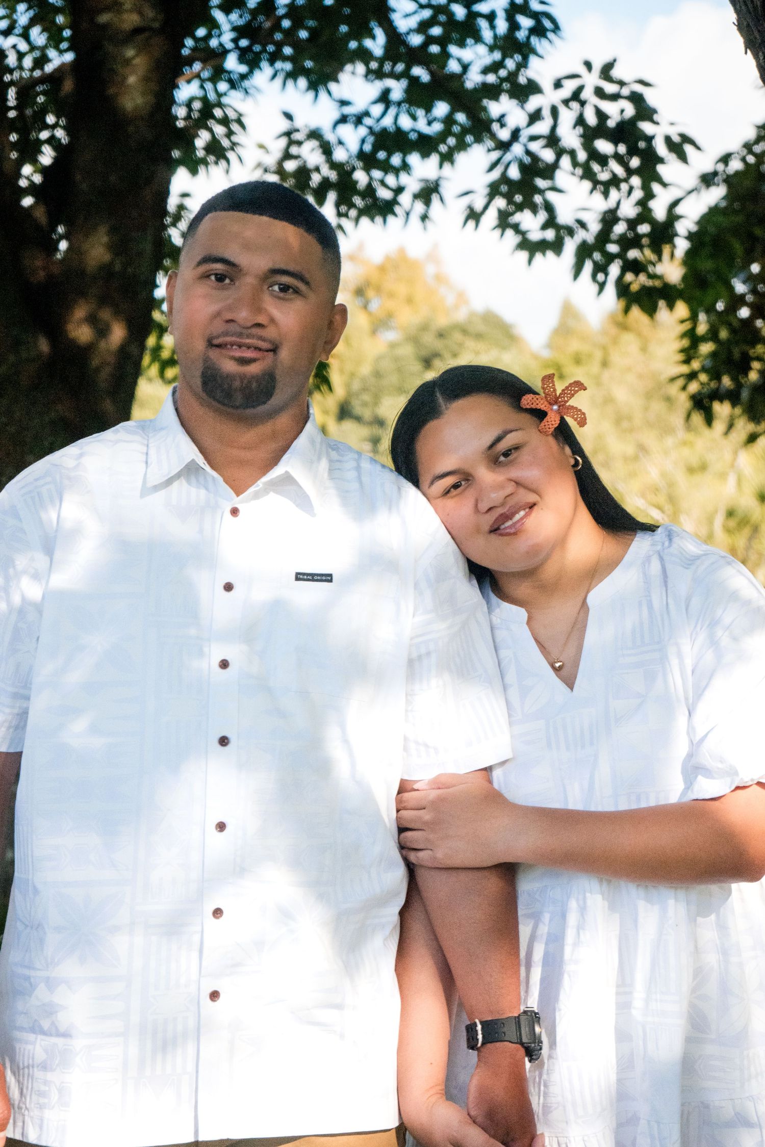 A couple in matching white outfits pose together outdoors with lush green trees in the background.
