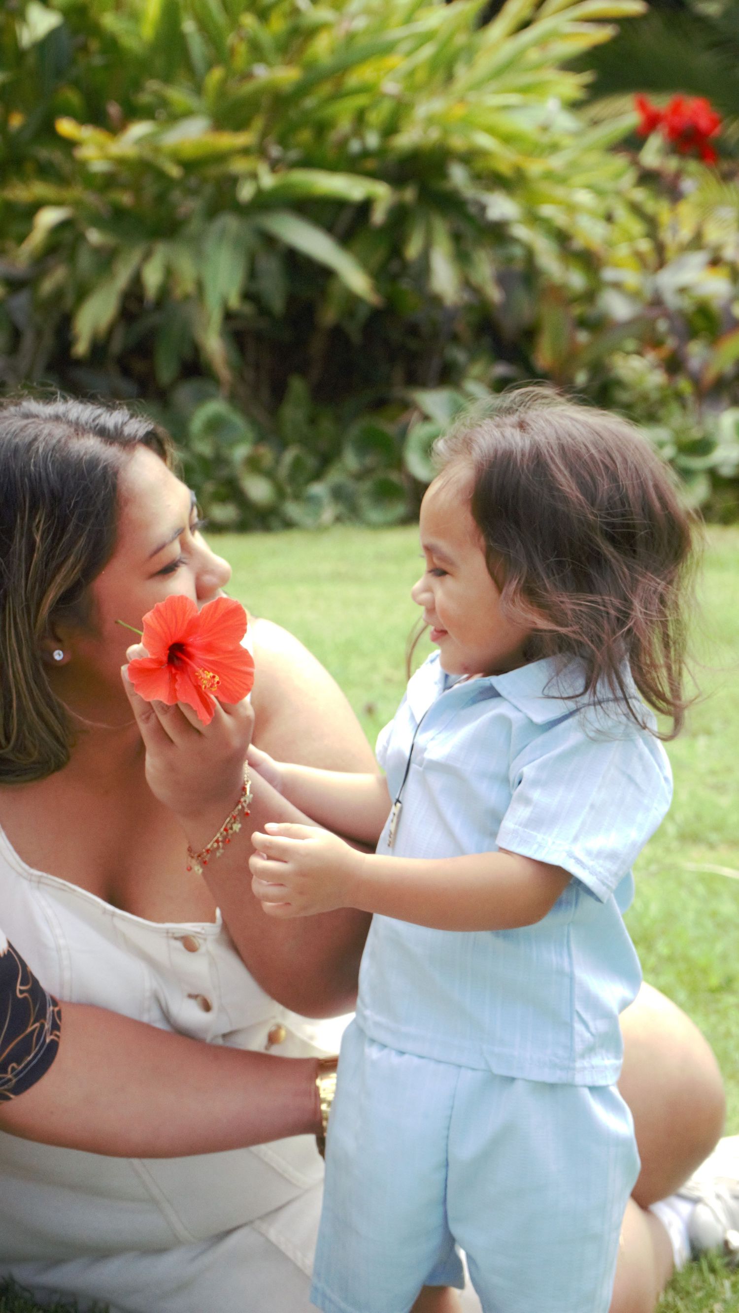 A mother and toddler share a sweet moment with a red hibiscus flower in a garden.