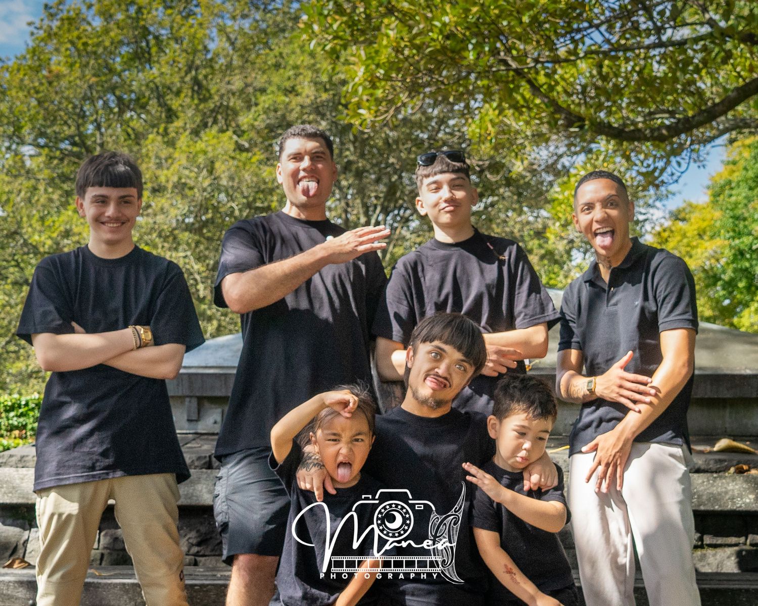 Group of seven males of various ages making fun poses together outdoors near a stone wall.