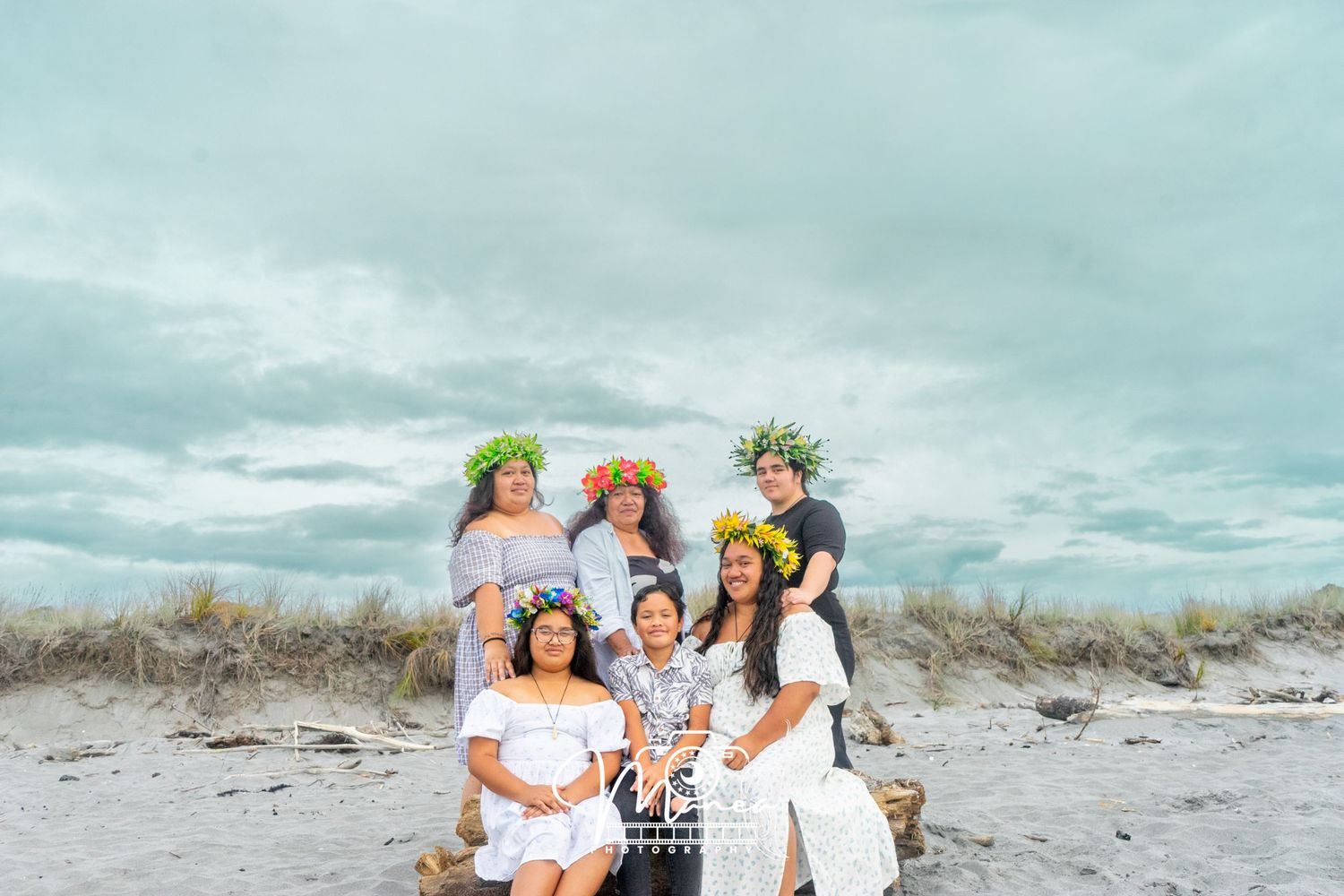 A group of six people pose together on a sandy beach, several wearing colorful floral crowns.