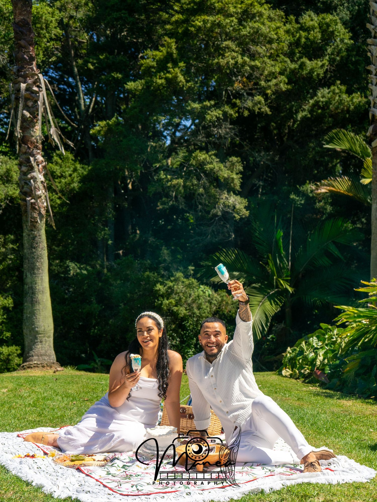Joyful couple in white outfits celebrating outdoors on picnic blanket in lush garden.