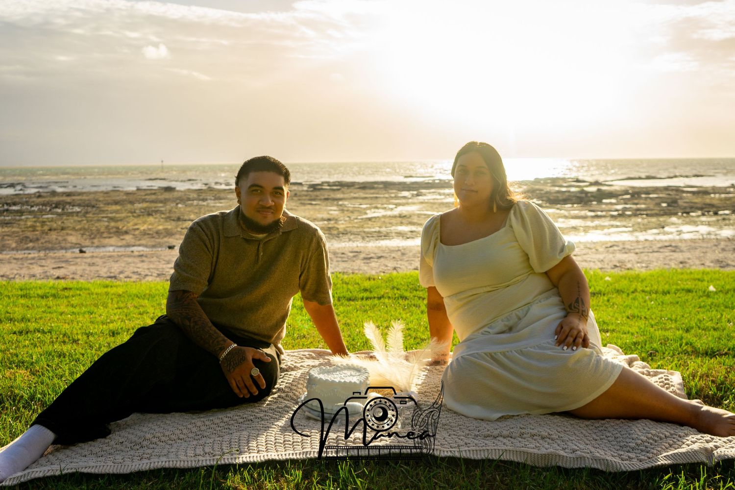 Couple sitting together on picnic blanket at sunset with ocean in background.