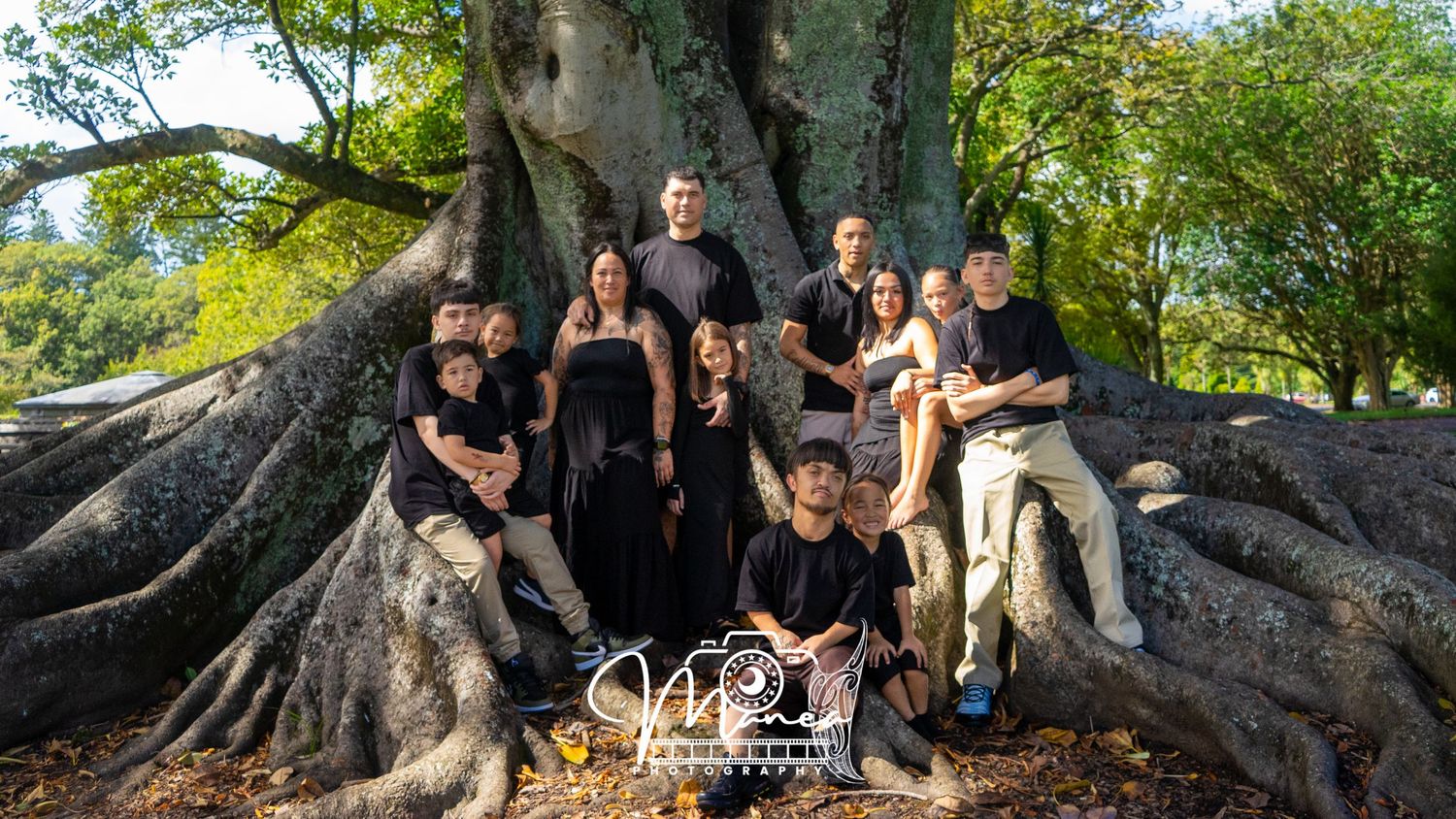 Large family group photo posed in front of a massive tree with sprawling roots outdoors.