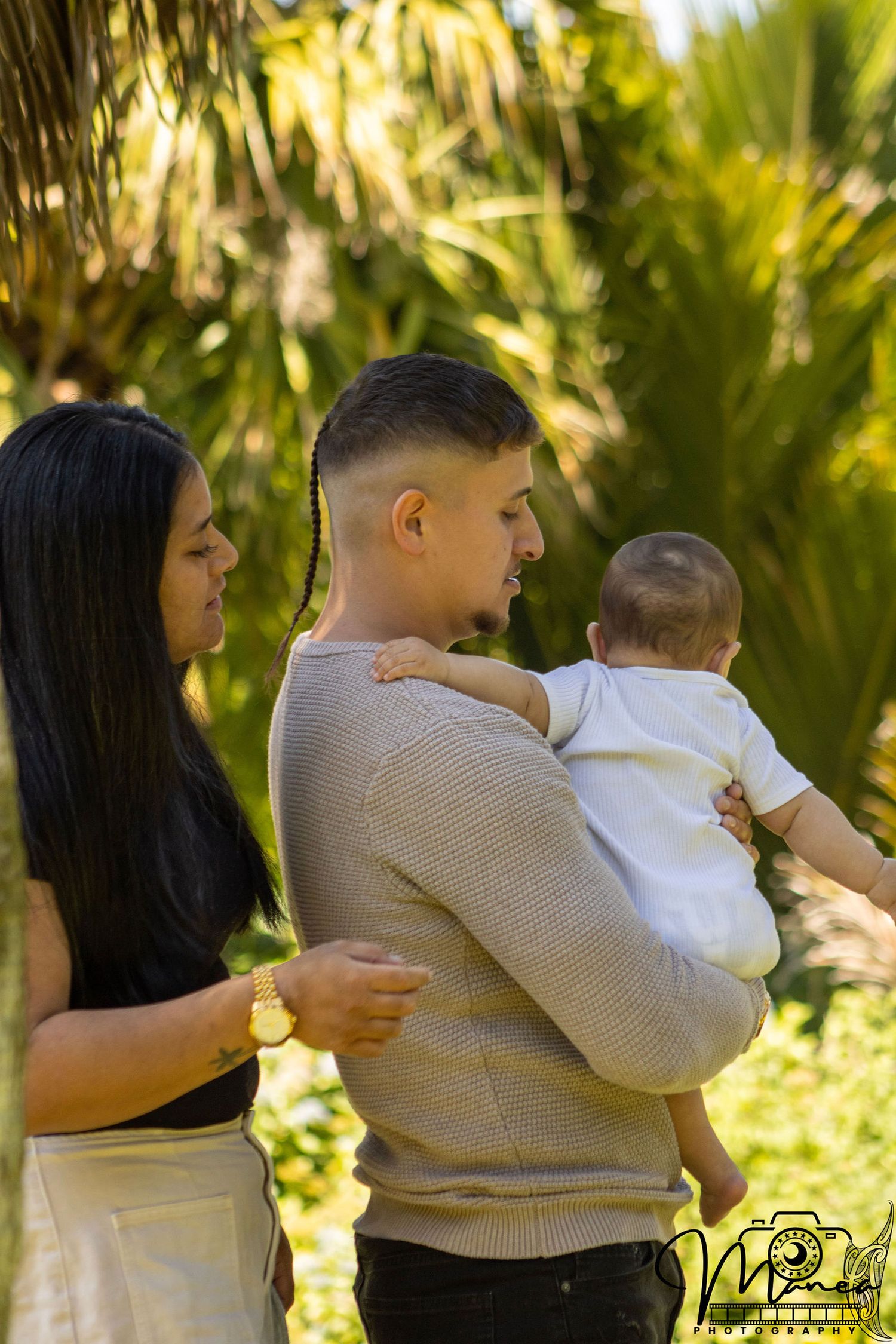 A couple admires their newborn baby outdoors surrounded by golden tropical palm trees.