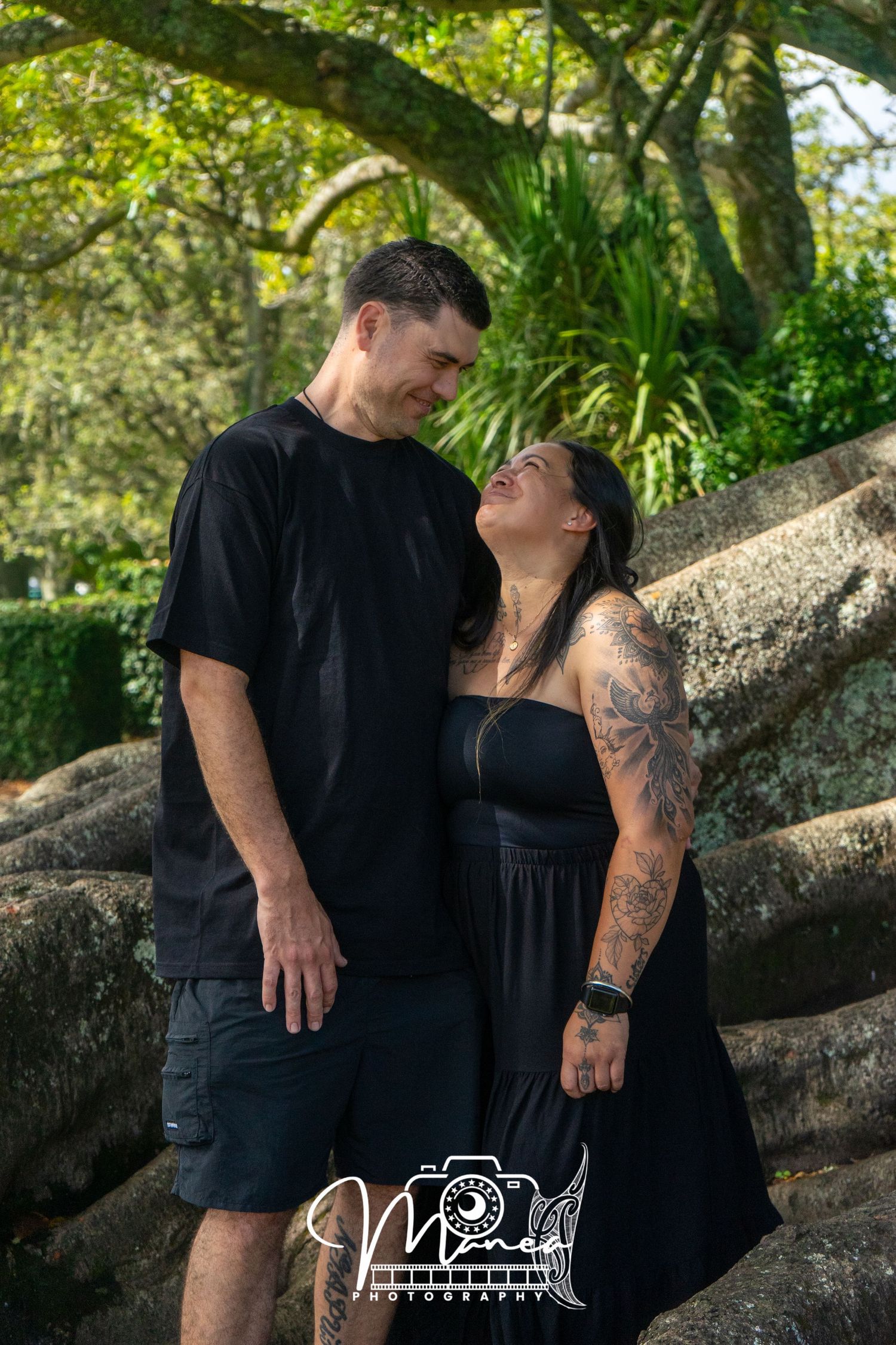 Tall man and tattooed woman in black outfits sharing a loving glance near large tree roots.