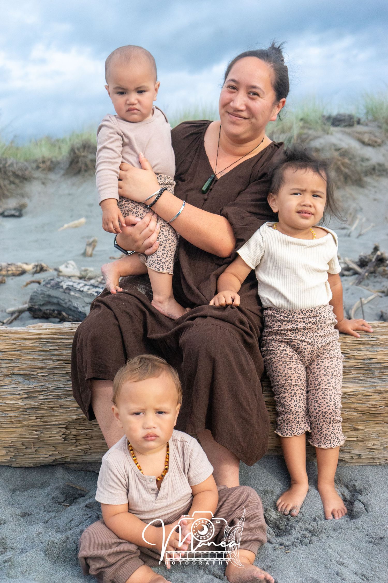 A mother sits on driftwood at the beach holding two toddlers while a third child sits in the sand below.