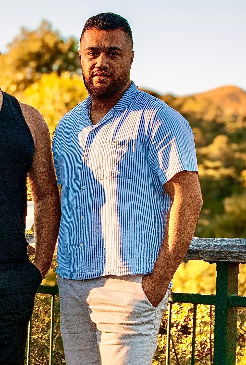 A man in a blue and white striped short-sleeve shirt stands outdoors with scenic hills behind him.