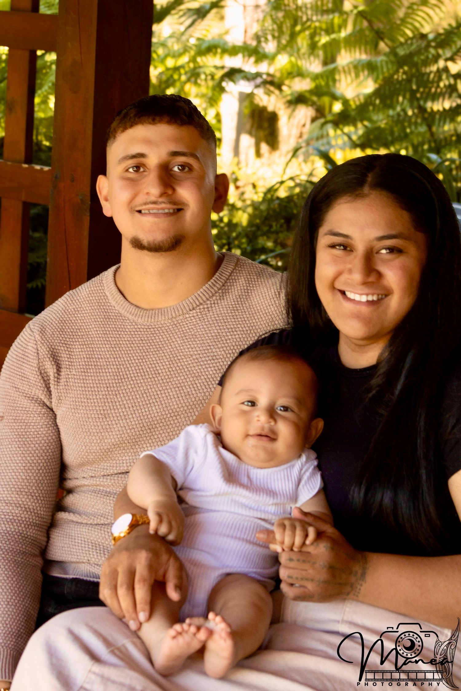 A young couple sits on a porch smiling while holding their baby between them.