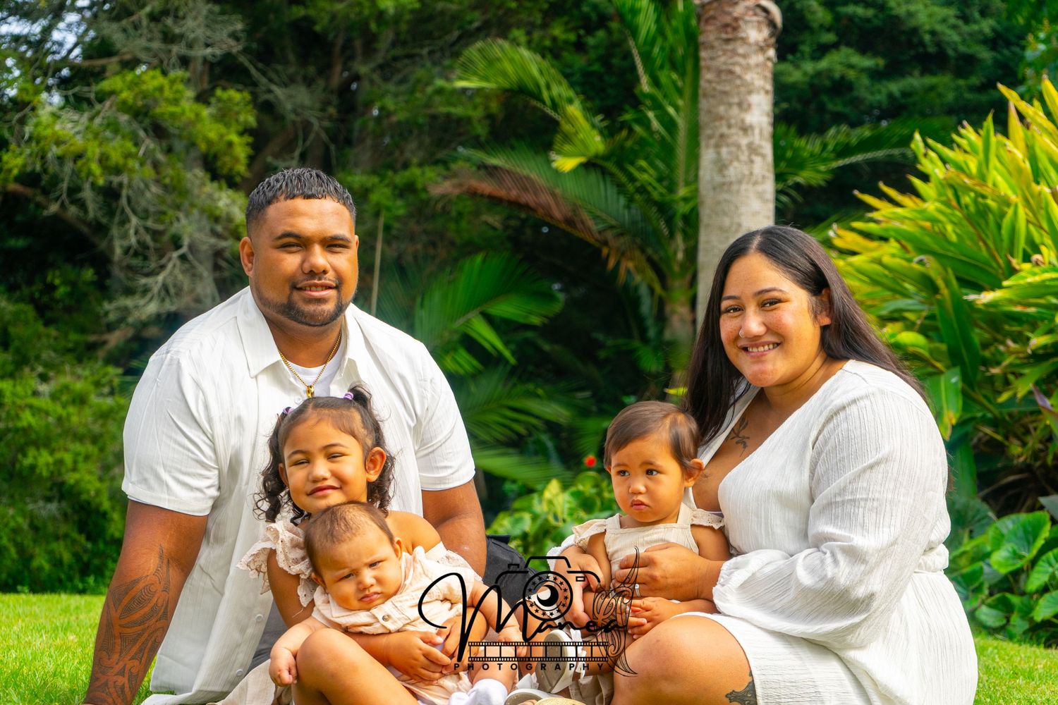 A family of four dressed in white poses outdoors on green grass with lush tropical plants behind them.