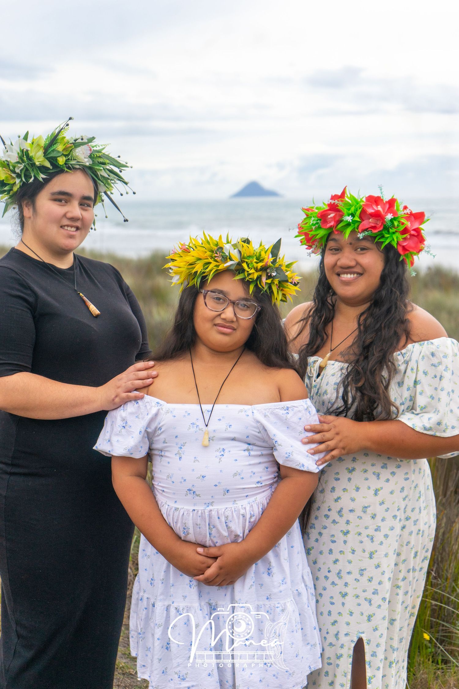 Three young women pose outdoors near the ocean, two wearing vibrant floral crowns and one in yellow.
