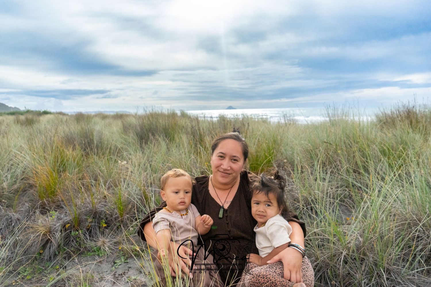 A grandmother sits with two toddlers in coastal sand dunes with tall grass and cloudy sky.