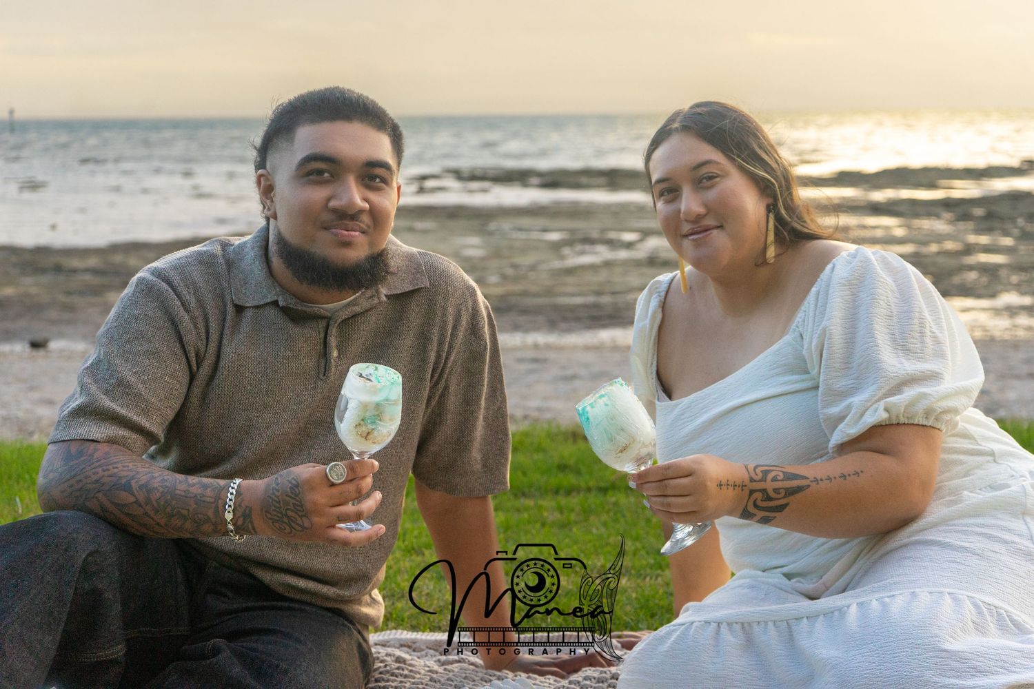 Smiling couple sitting on blanket by ocean at sunset holding wine glasses.
