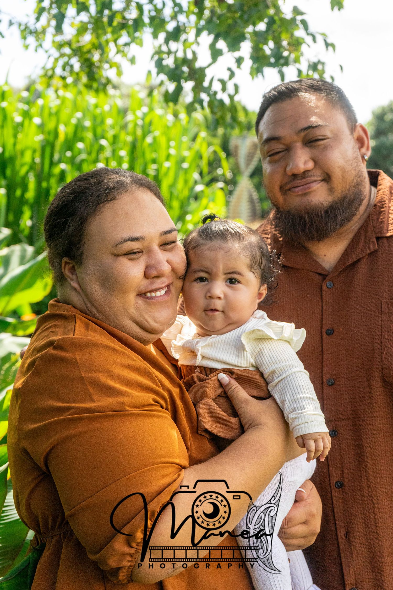 A young family with a baby girl poses outdoors surrounded by lush green plants.