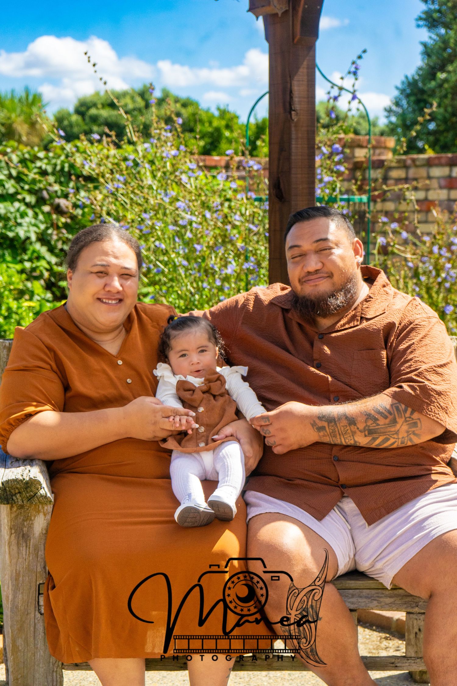 Pacific Island family sitting together on a bench in a sunny garden with colorful flowers.