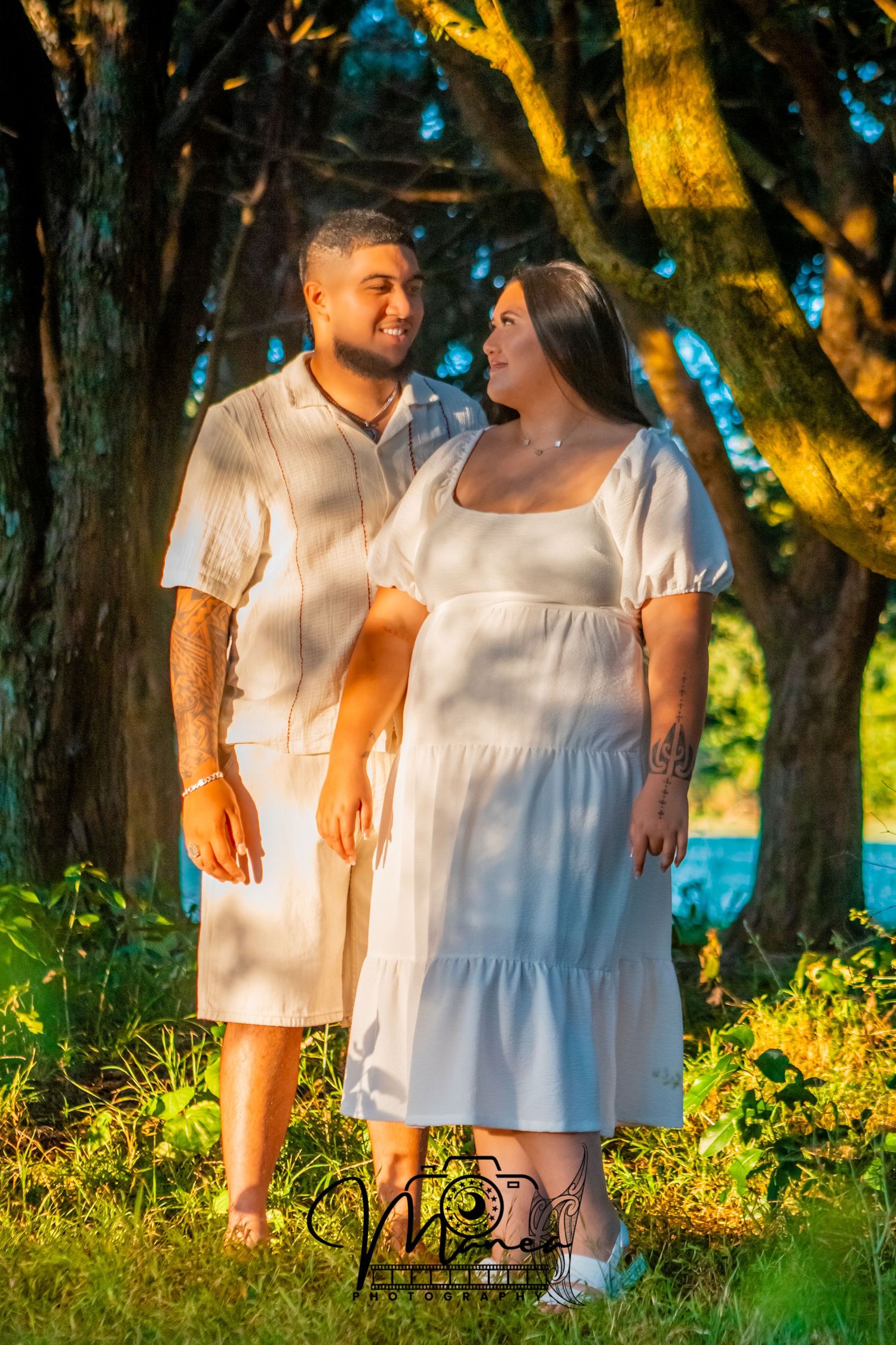 Expecting couple in white outfits standing together in a sunlit wooded area, smiling at each other.