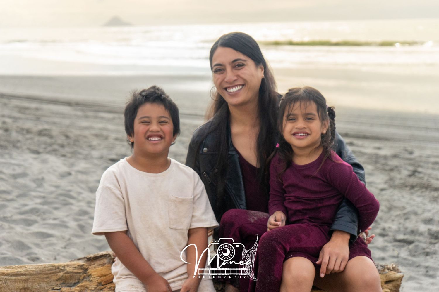 Smiling woman sitting on driftwood with two young children on a beach at sunset.
