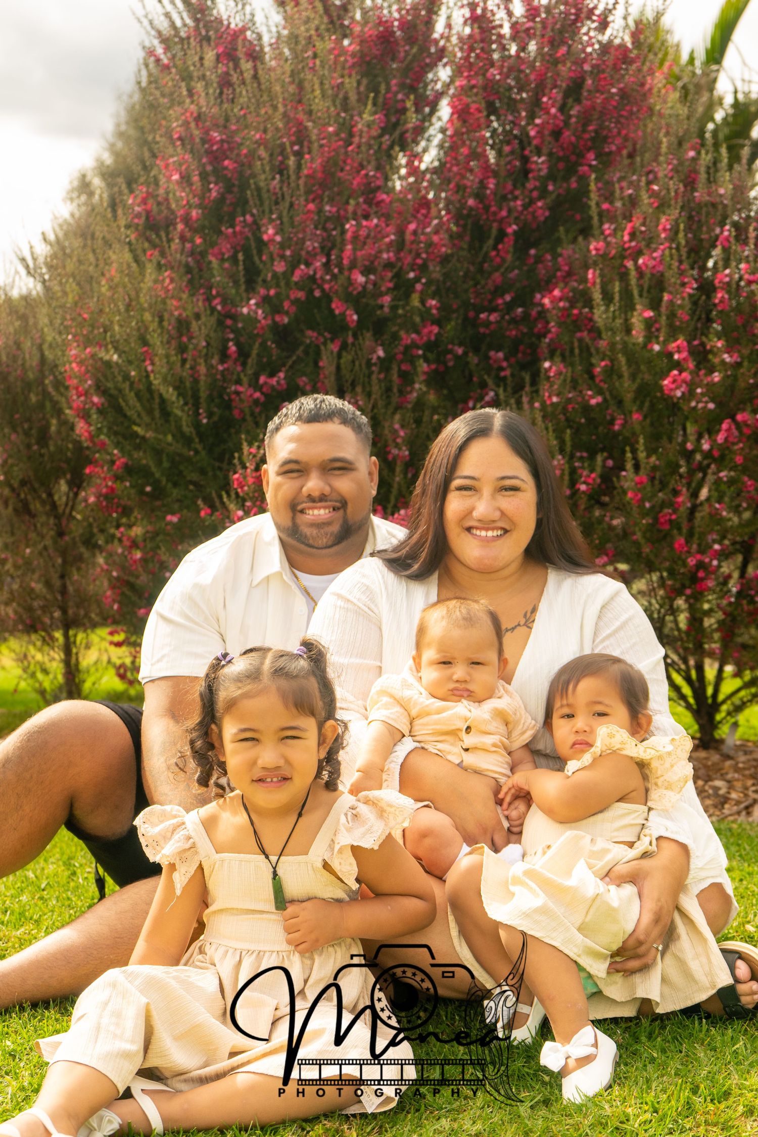 A family of five sits on green grass in front of blooming red trees during an outdoor photo session.