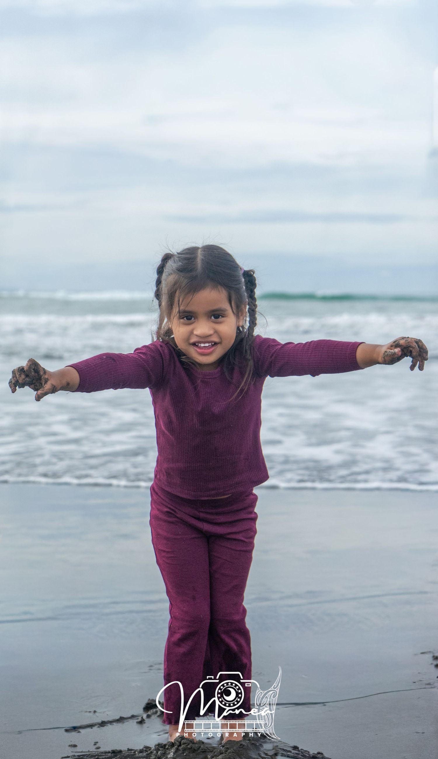 A joyful young girl in a maroon outfit spreads her arms wide on a beach, smiling with sandy hands near ocean waves.