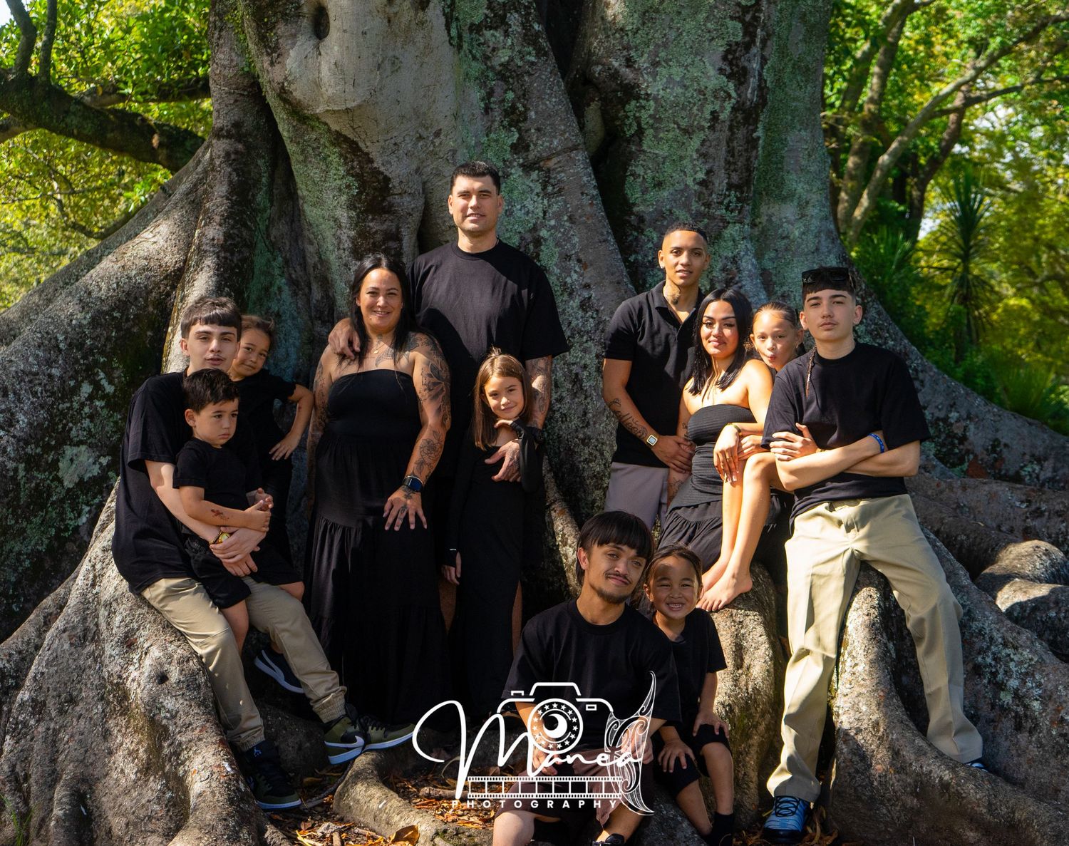 Extended family portrait of eleven people gathered around large tree roots in a park.