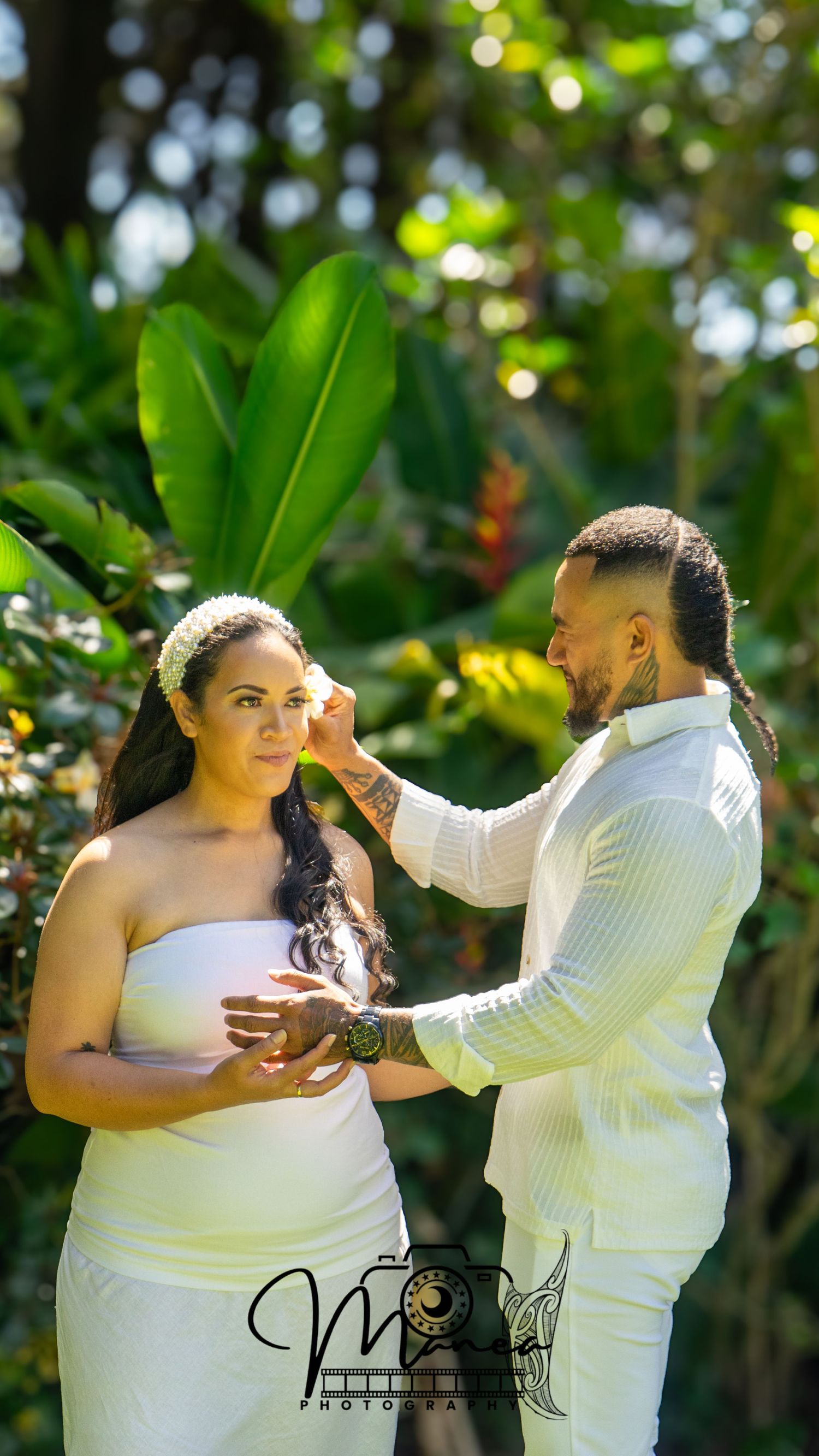 Couple in white outfits sharing tender moment surrounded by lush tropical greenery.