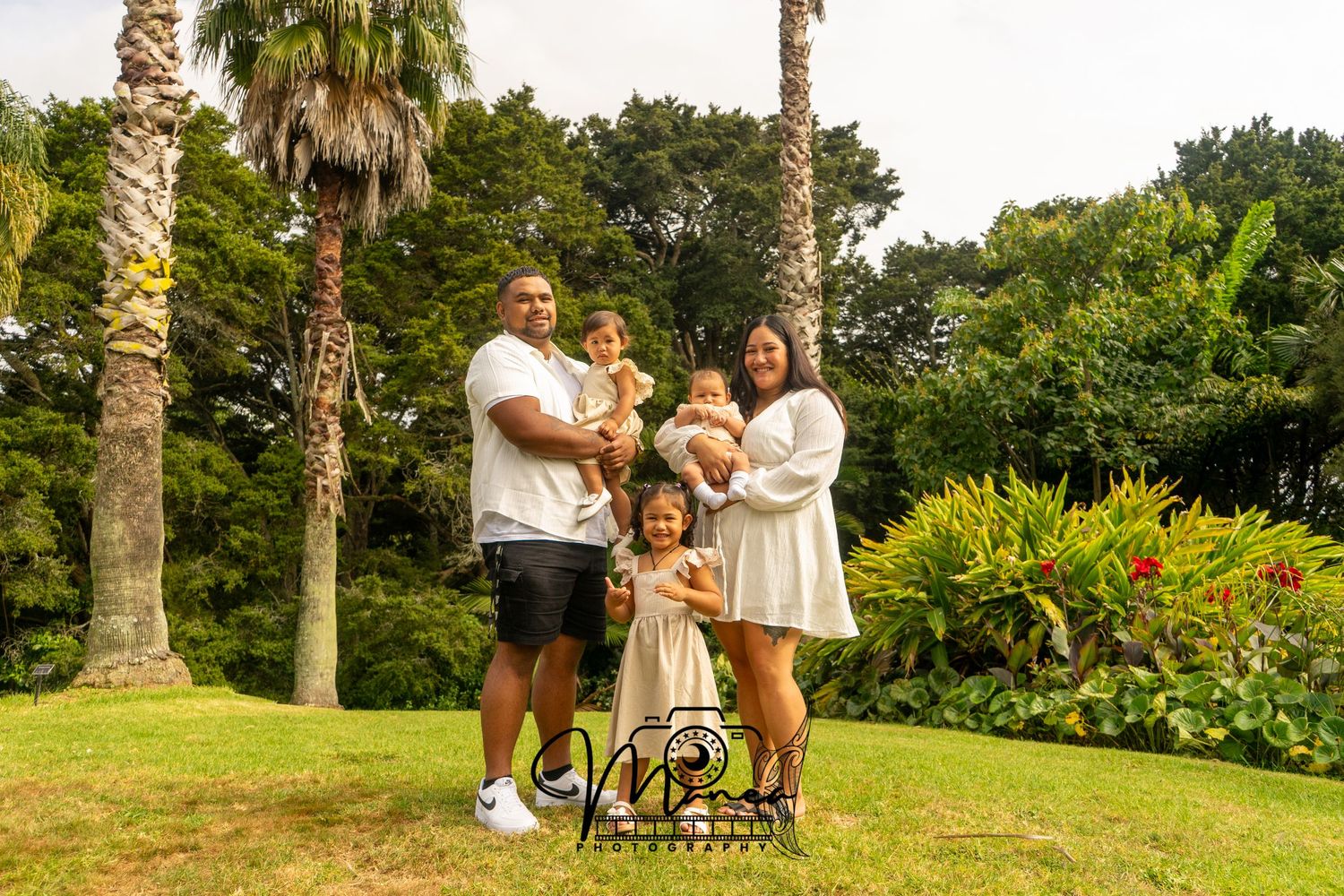 Happy family of five posing outdoors on green lawn with tropical palm trees in background.