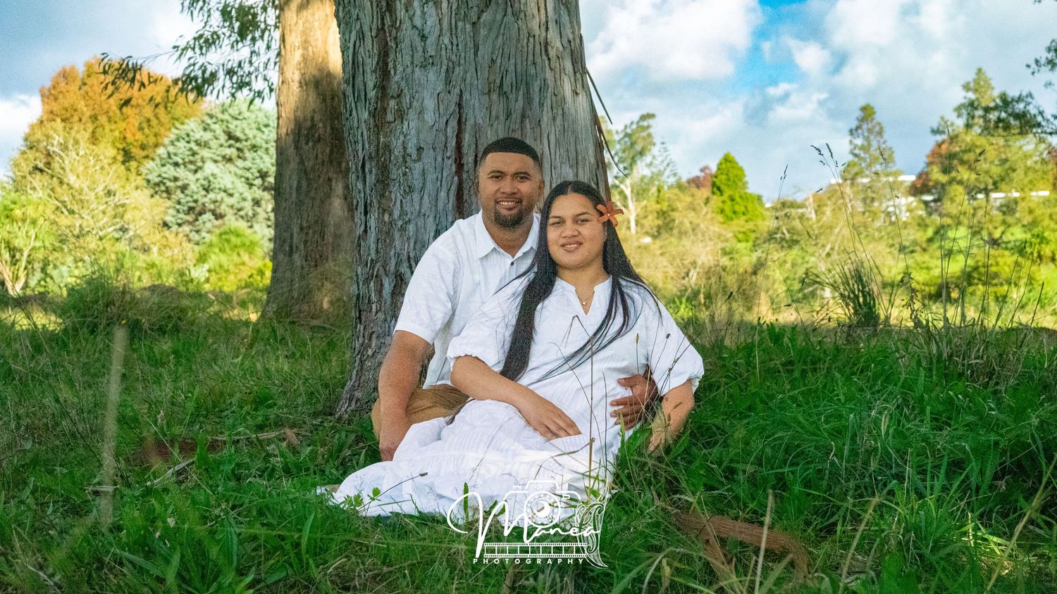 Couple in matching white outfits sitting together at the base of a large tree in a scenic park.