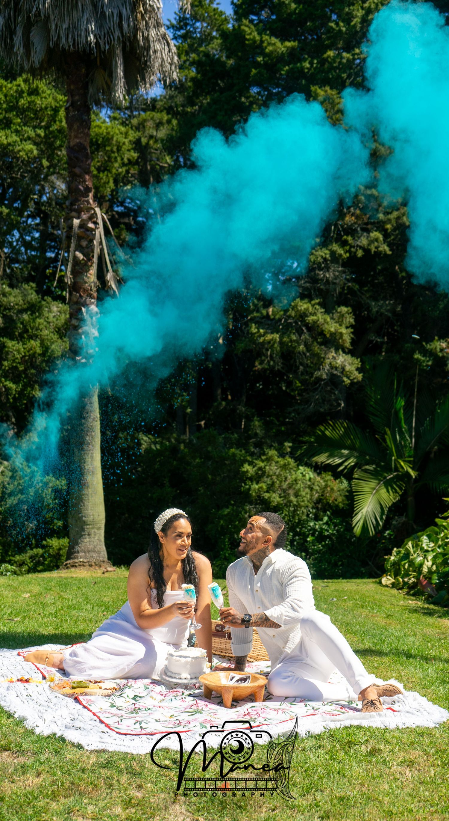 Couple in white sitting on blanket outdoors with teal gender reveal smoke bomb.