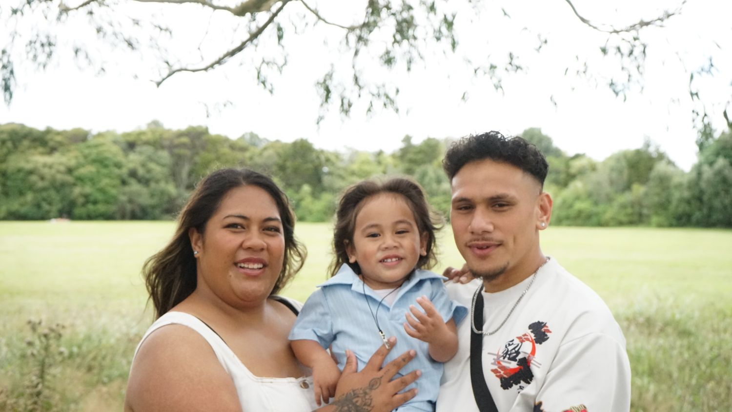 Smiling young family of three posing together in an open green park with trees behind them.