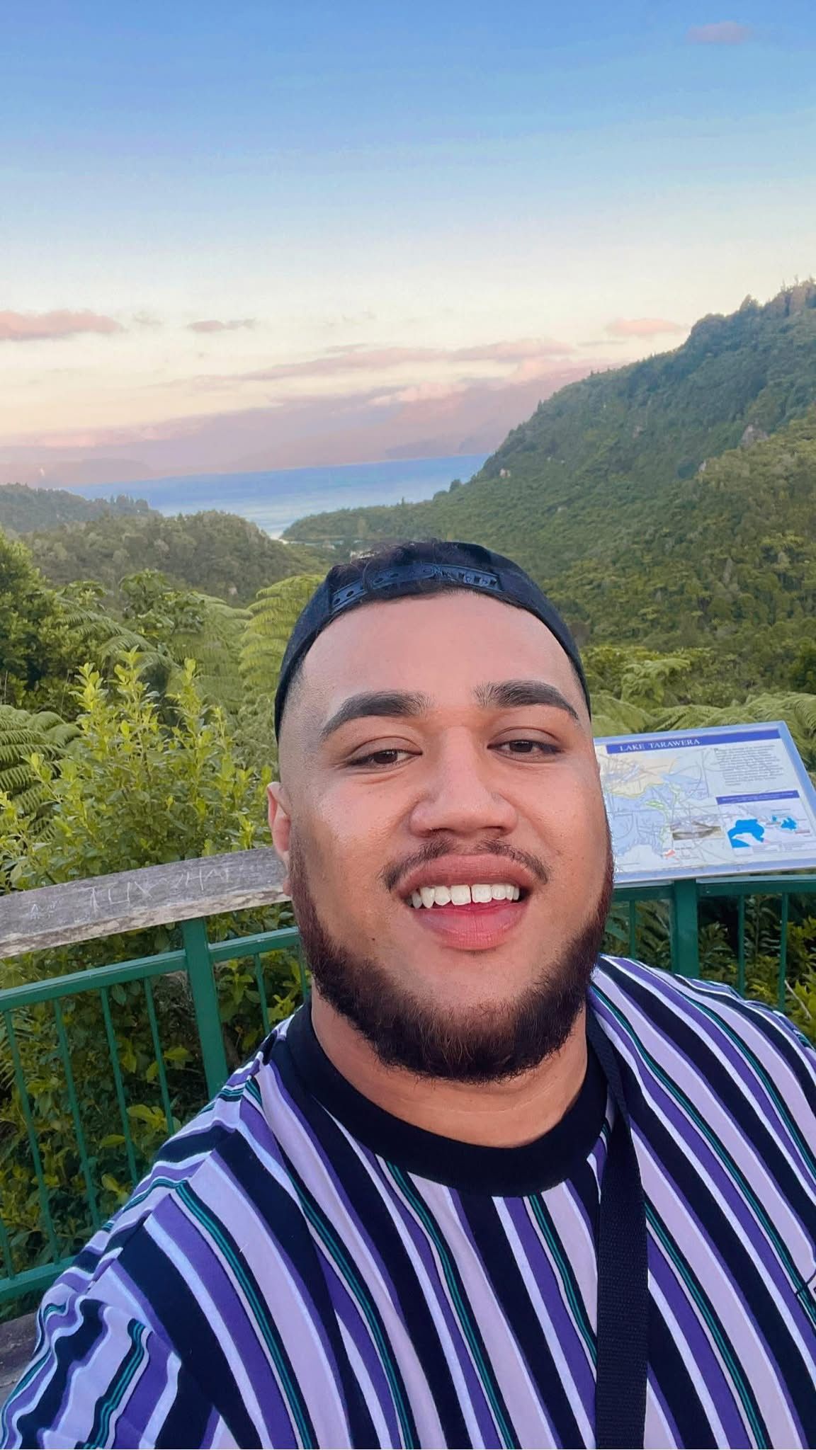 A man smiles for a selfie at a scenic mountain valley overlook under a colorful sky.