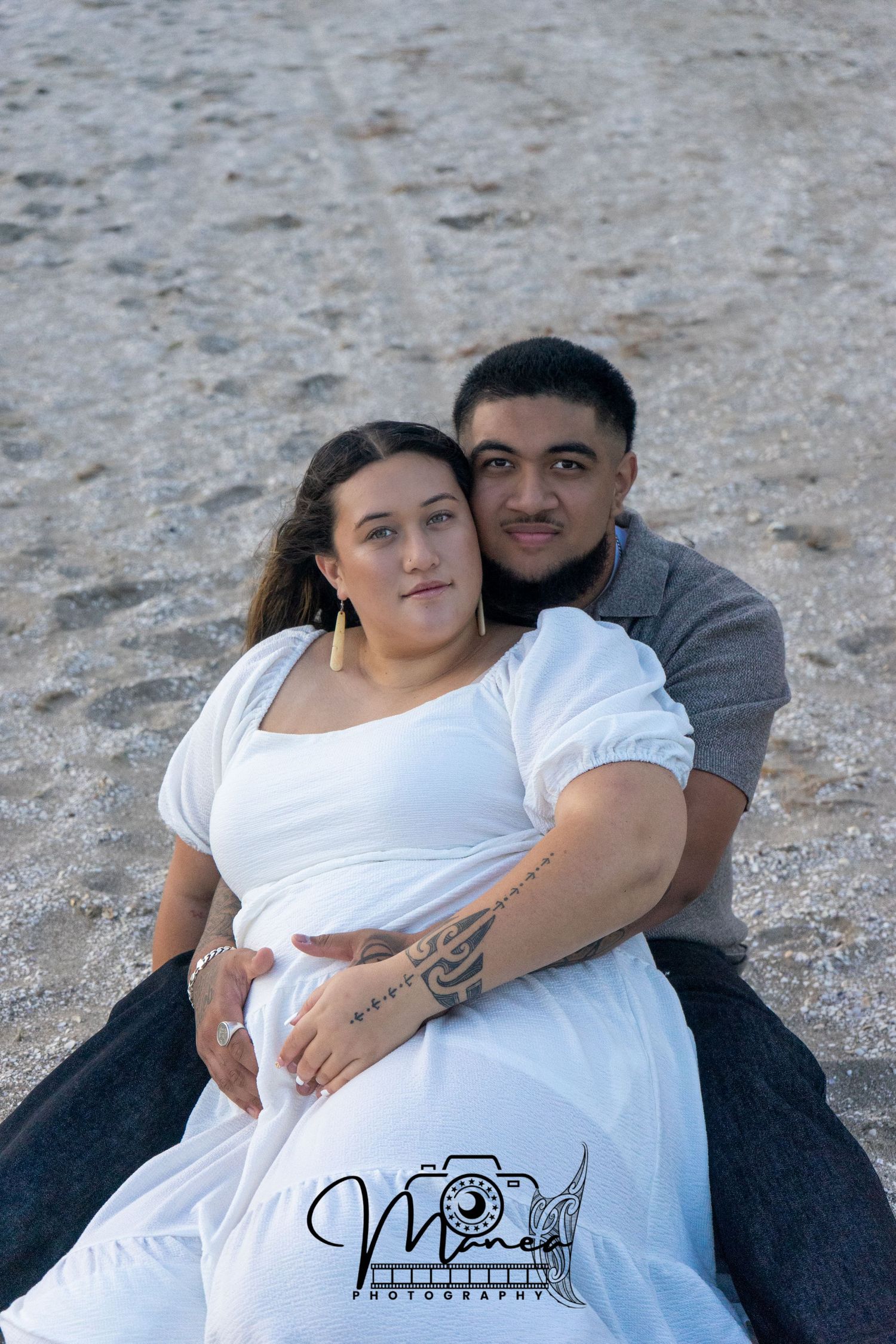 Pregnant couple sitting on beach sand, woman in white dress with partner embracing her baby bump.