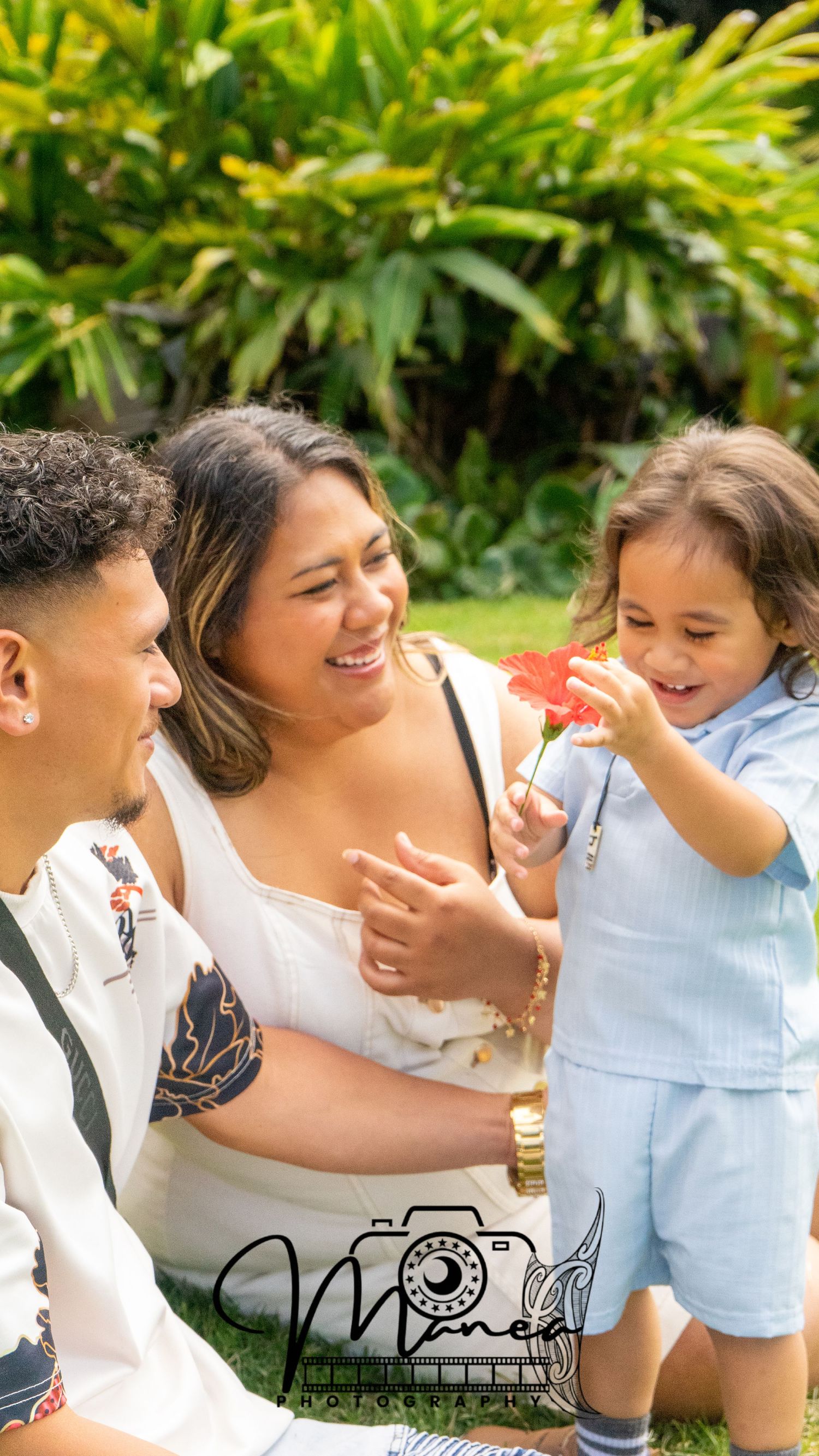 Young family sitting on grass as toddler son holds a bright red flower in sunny garden.