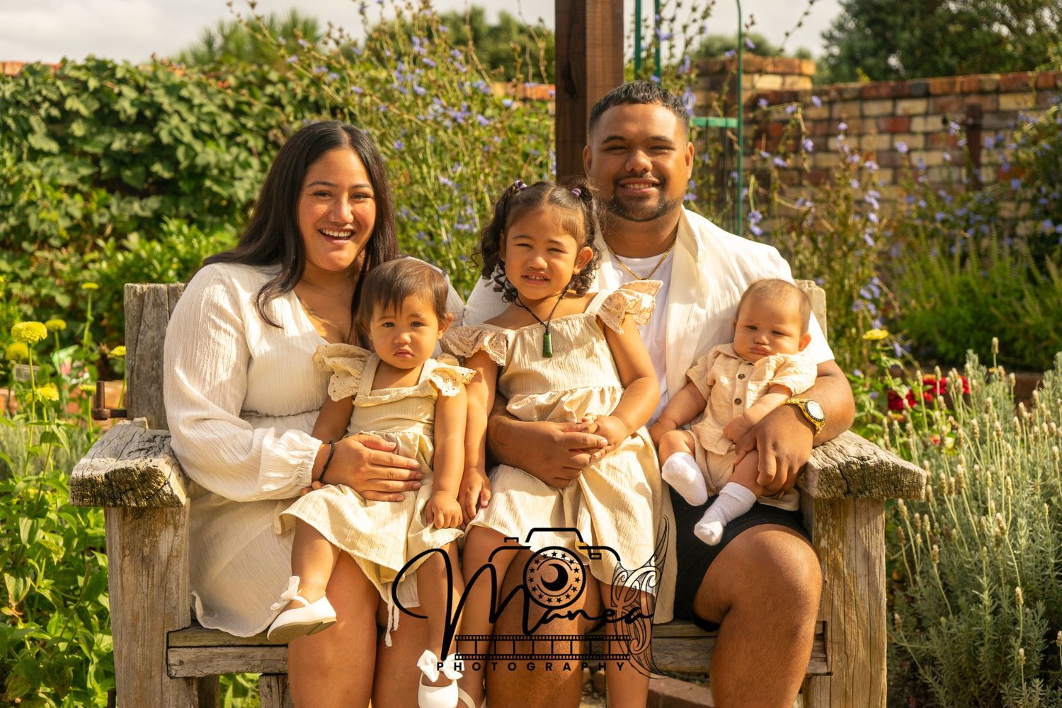 A family of five in matching white and gold outfits poses on a wooden bench in a garden setting.