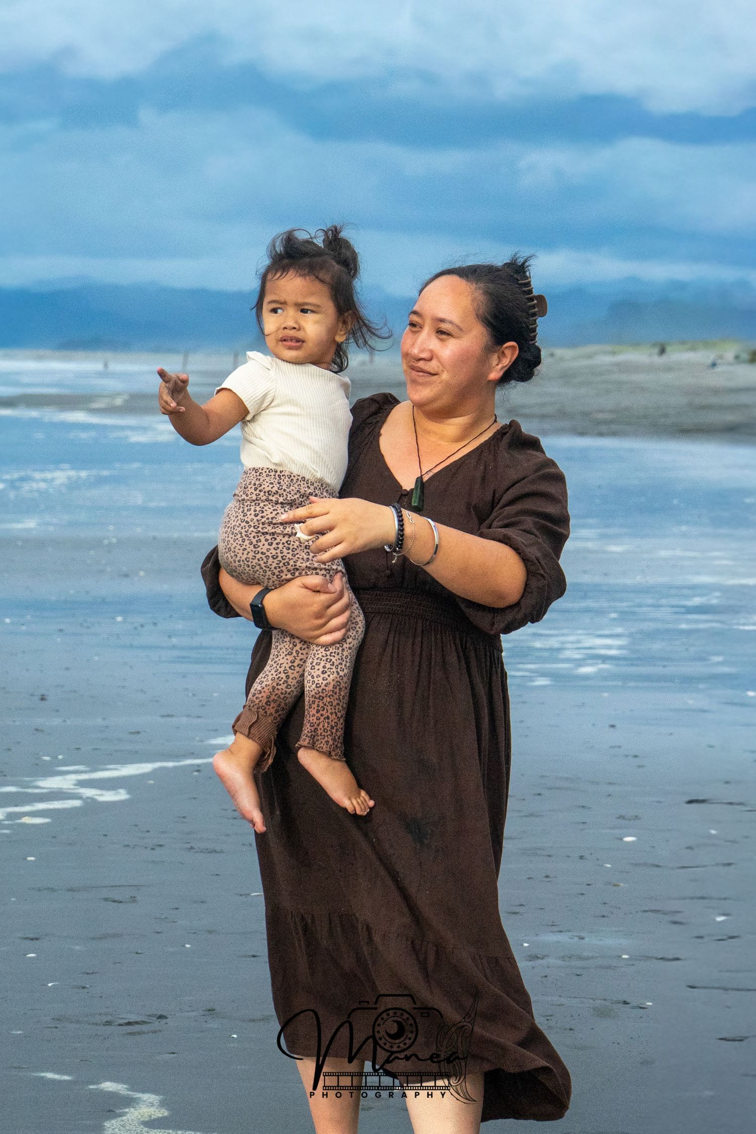 Mother in brown dress holding her smiling toddler on a cloudy beach shoreline.