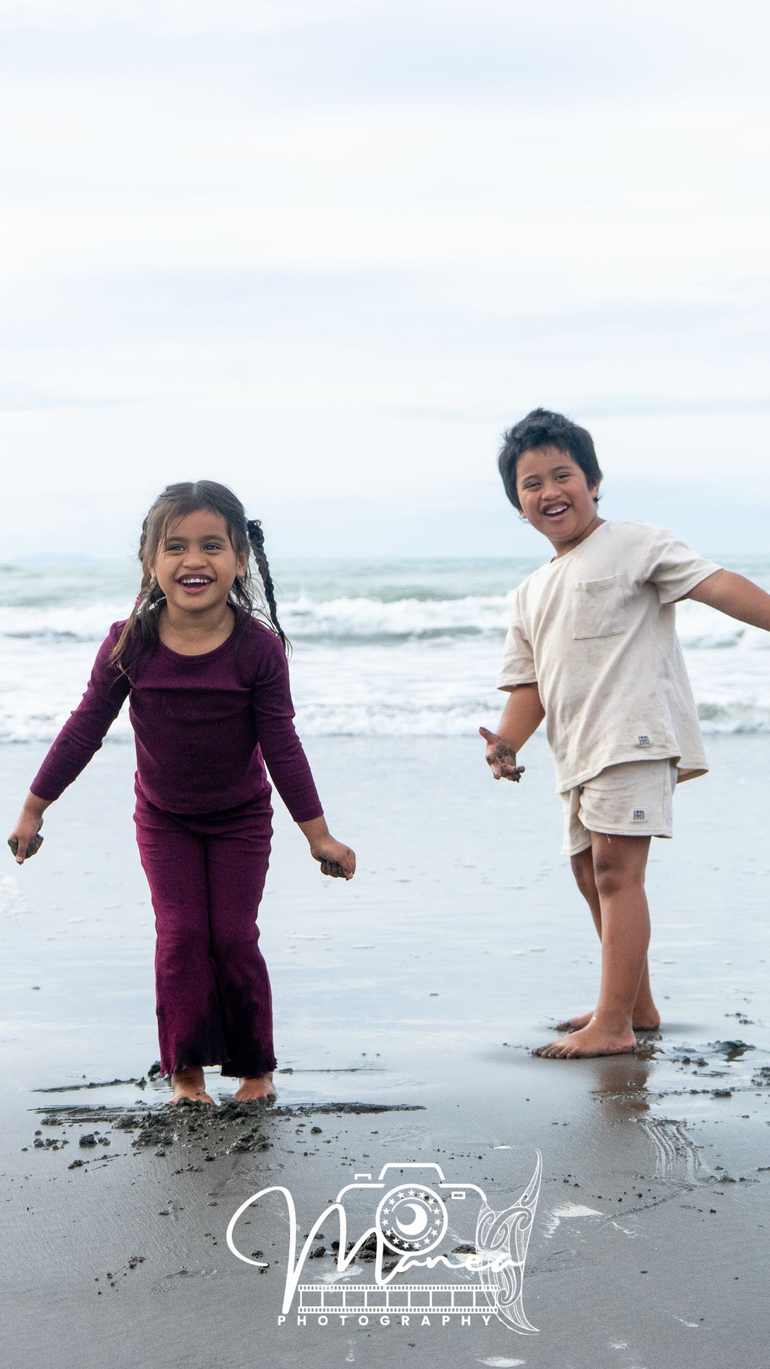 Two young children laughing and playing together on a sandy beach near the ocean waves.