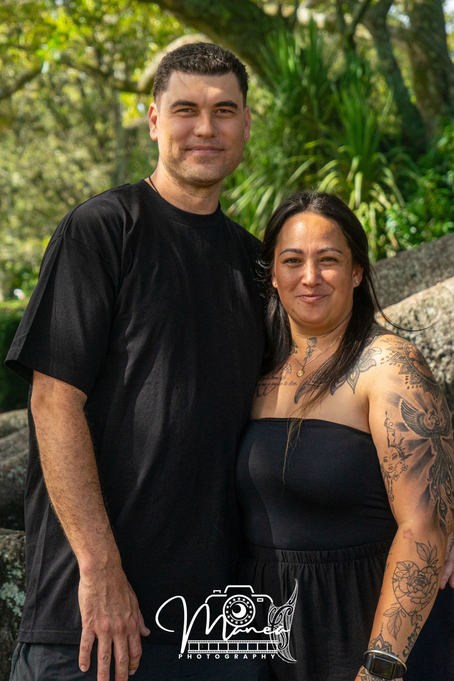Couple in matching black outfits posing together in a lush green garden setting.