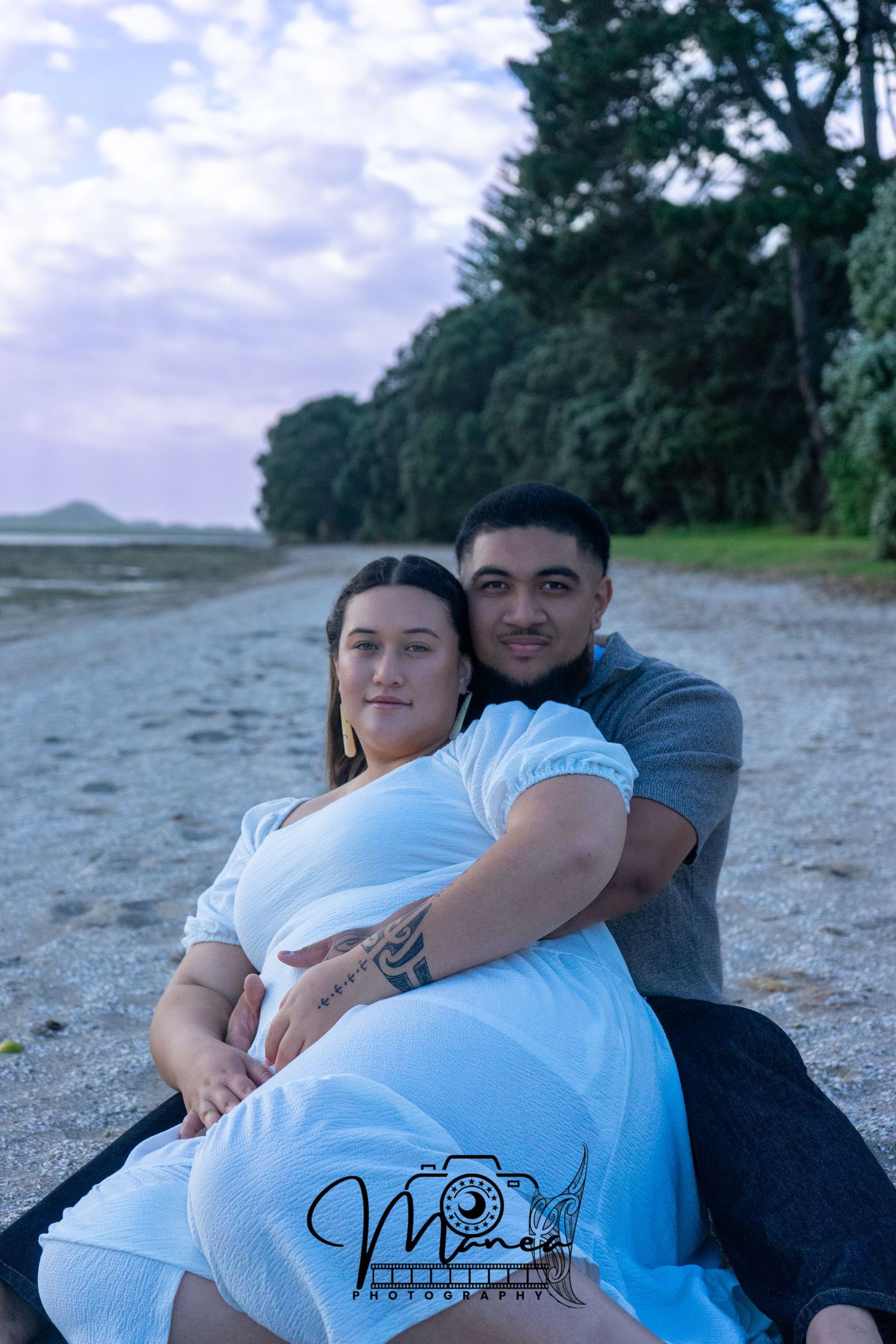 Pregnant woman in white dress sitting on a beach with her partner embracing her from behind.