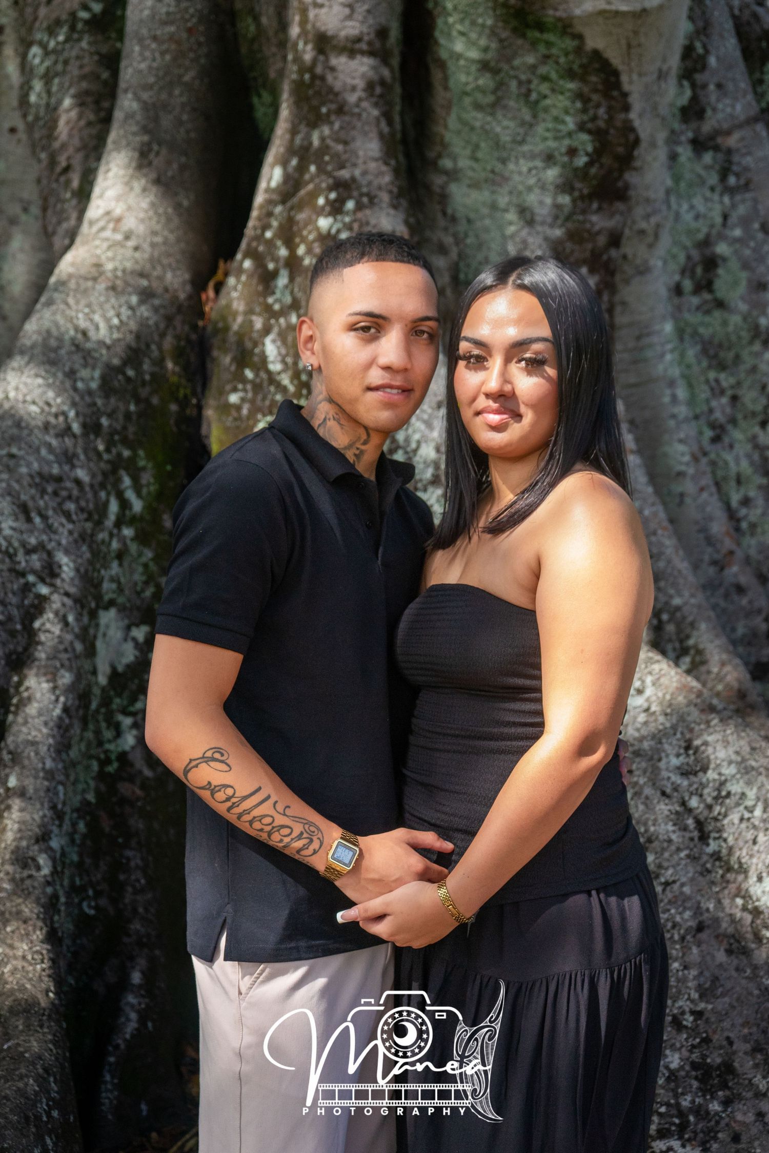 Young couple posing in front of large tree roots, man with neck tattoo in black polo shirt.