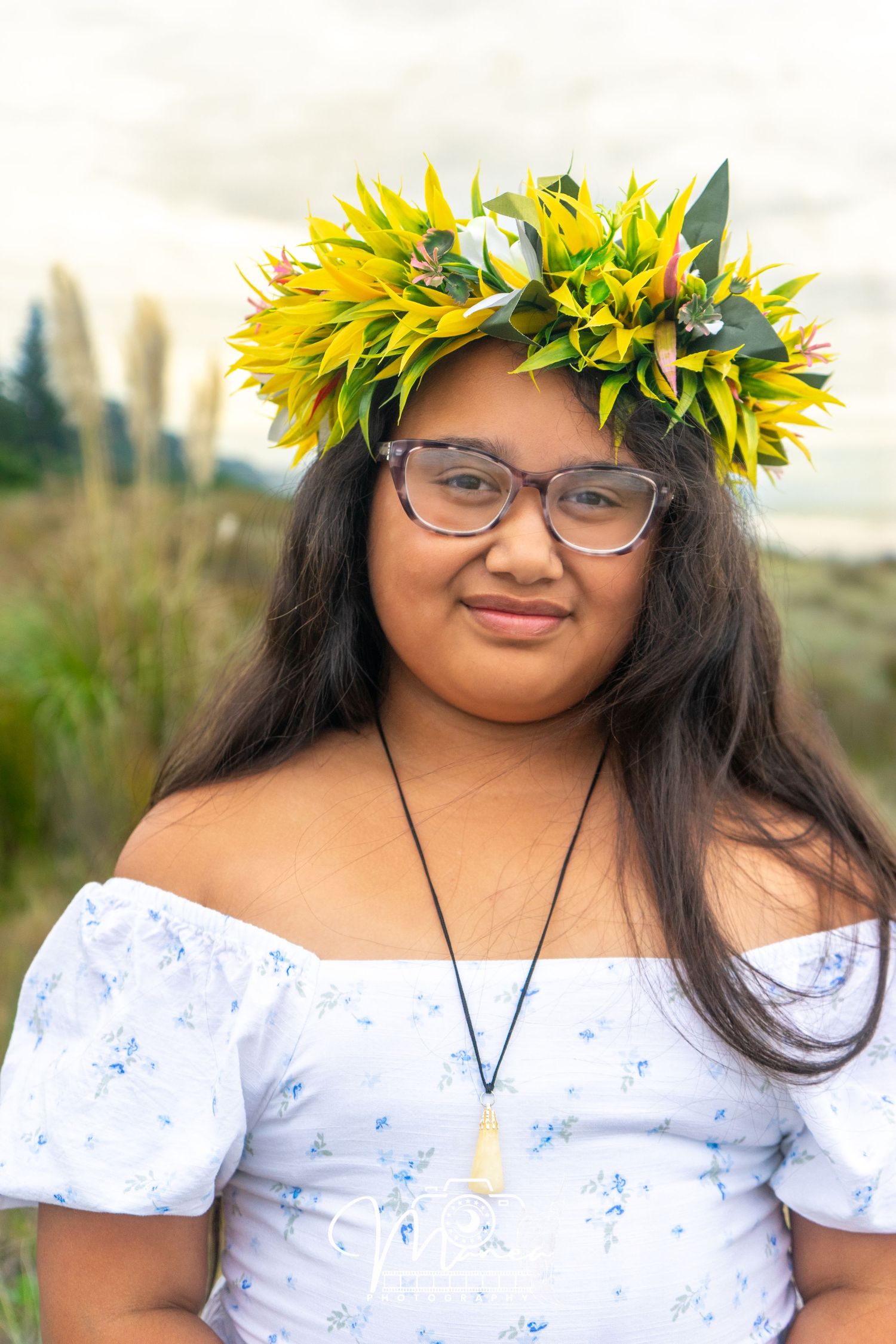 A young woman wearing a yellow floral crown and white off-shoulder dress poses outdoors smiling.