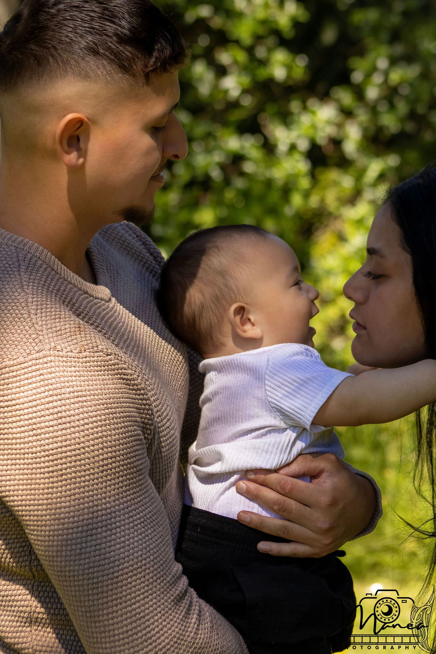 Loving parents holding their newborn baby outdoors surrounded by green foliage in warm sunlight.