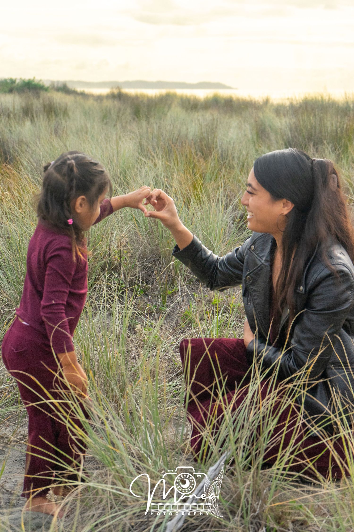 A woman crouches in beach dunes grass, smiling while interacting with a young girl in burgundy.